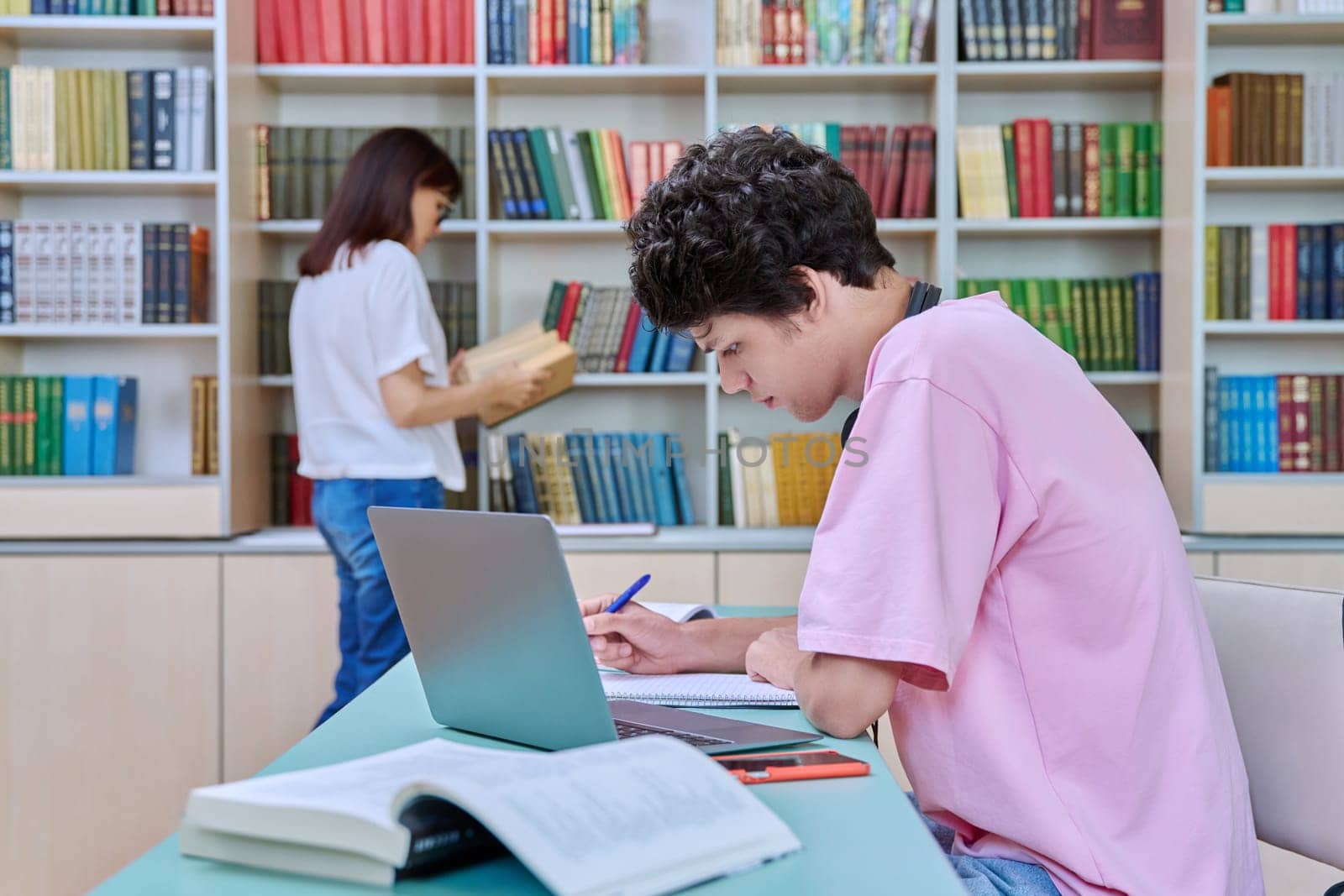 Young male student sitting with computer laptop books in college library. Guy 18-20 years old with headphones typing on laptop. Knowledge, education, youth, college university concept