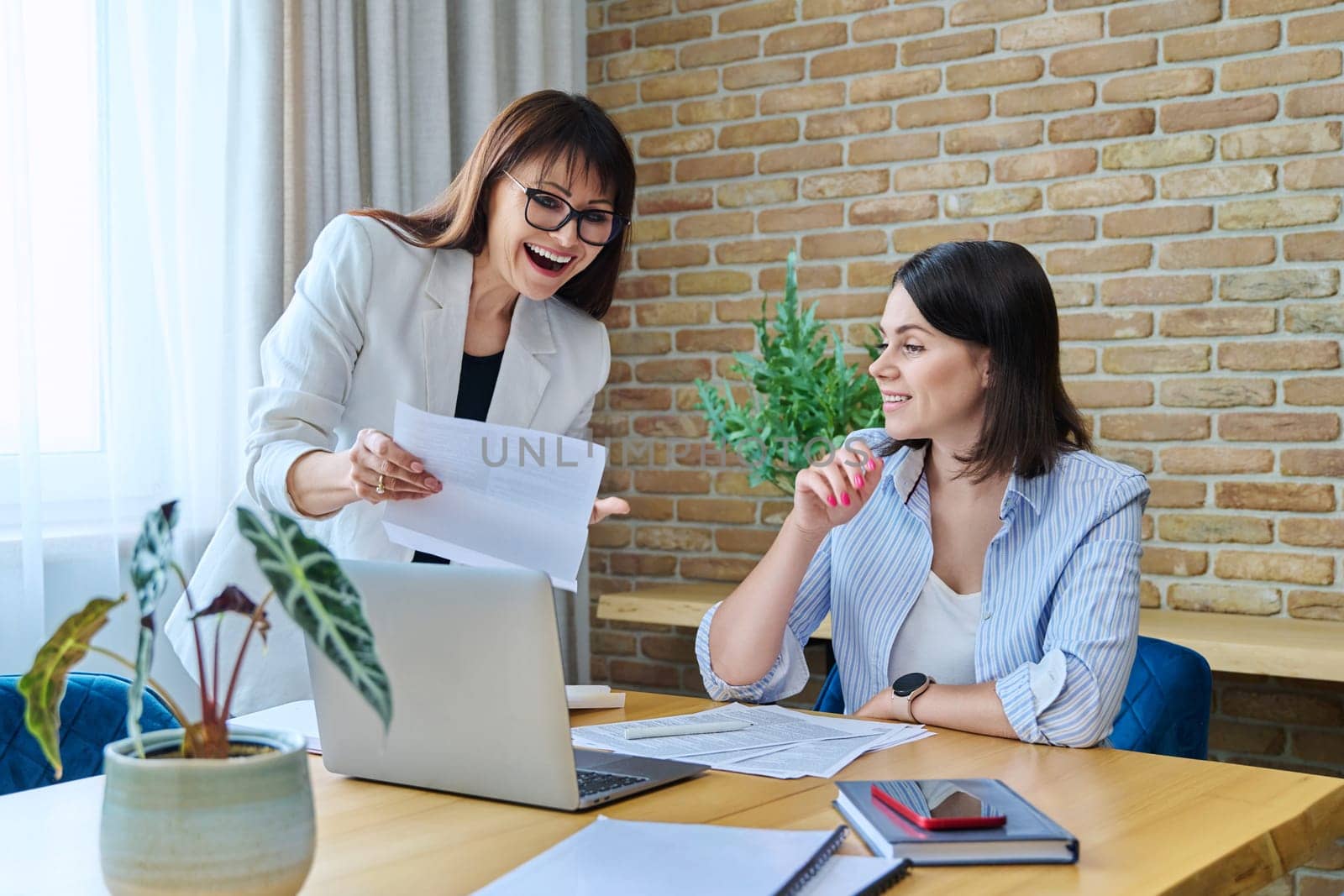 Two joyful happy smiling business women colleagues sitting at large table in office with business papers contracts. Business ceo work law finance mentoring consulting teamwork people job concept