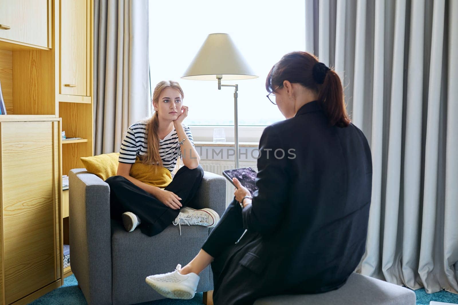 Young female teenager 16, 17 years old talking with counselor psychologist behavior social worker teacher, sitting in chair in office. Psychology psychotherapy therapy mental health adolescence youth