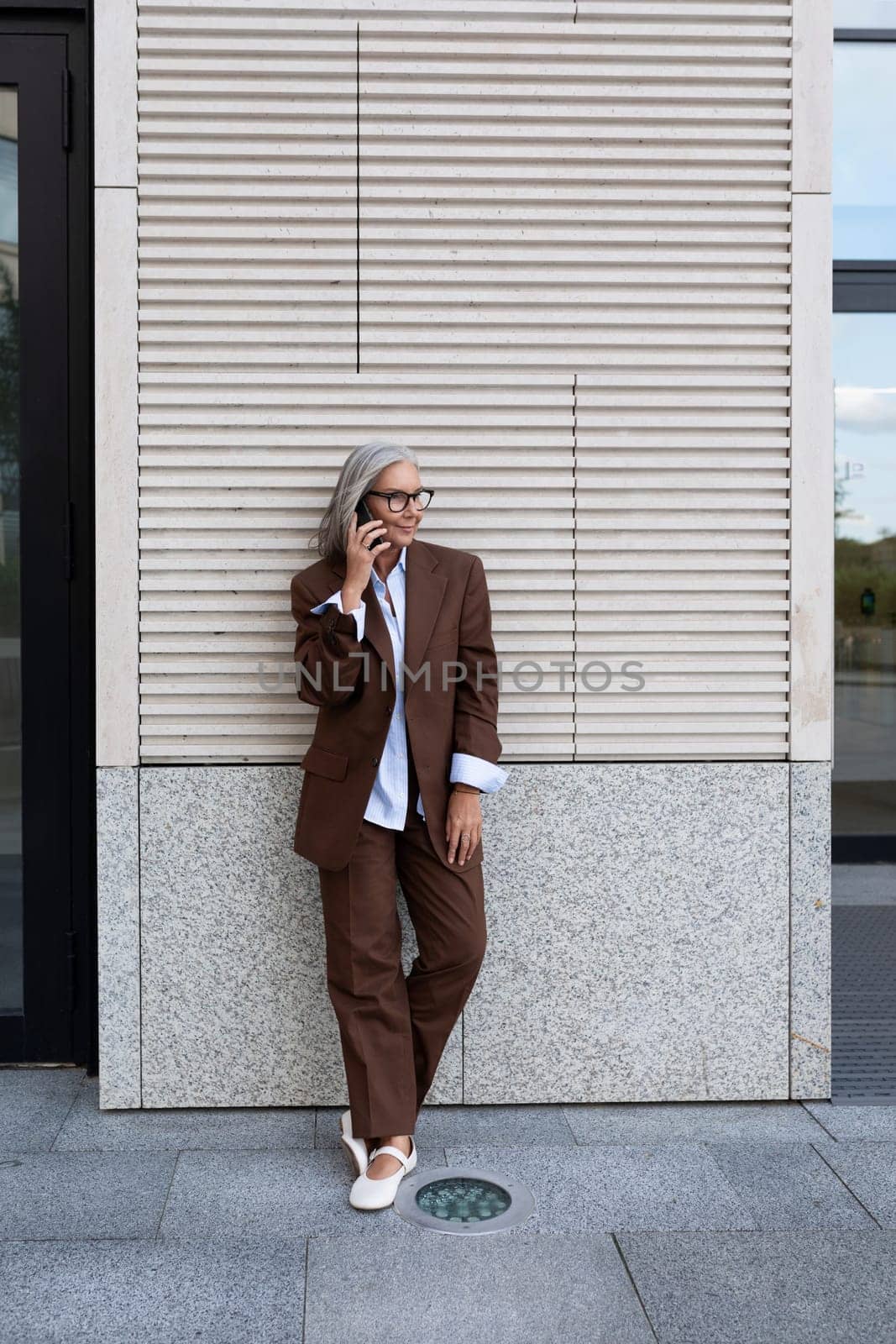 a successful gray-haired business grandmother dressed in a stylish brown suit stopped at the wall of the business center to use her smartphone.