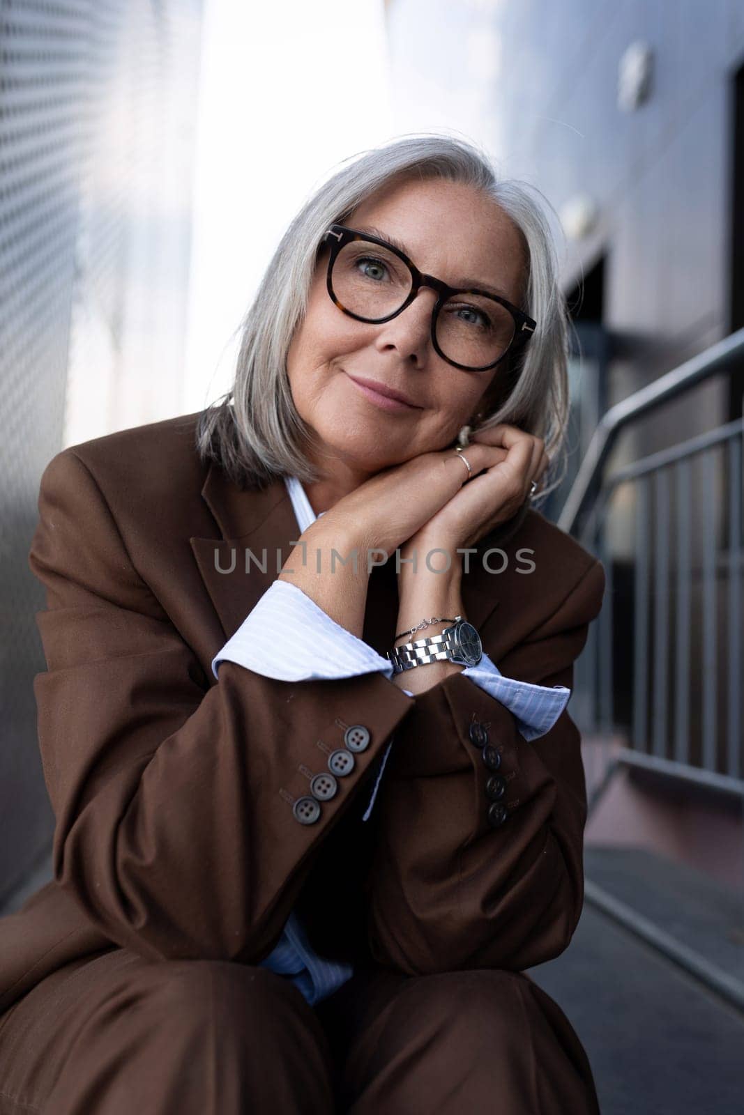 well-groomed healthy slender gray-haired mature business lady dressed in a stylish brown jacket sits against the backdrop of a business building.