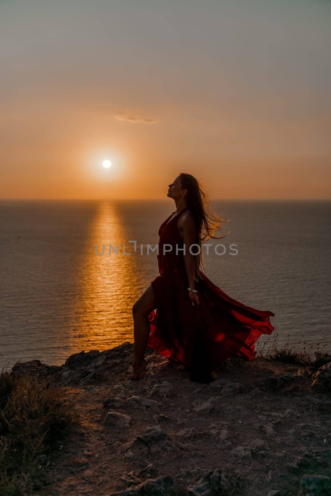 Woman sunset sea red dress, back view a happy beautiful sensual woman in a red long dress posing on a rock high above the sea on sunset