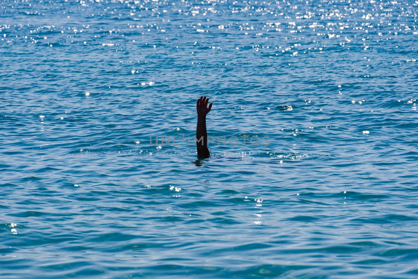 Drowning man concept, with a male raised hand with an open palm visible waving for help above a blue sea with low waves.