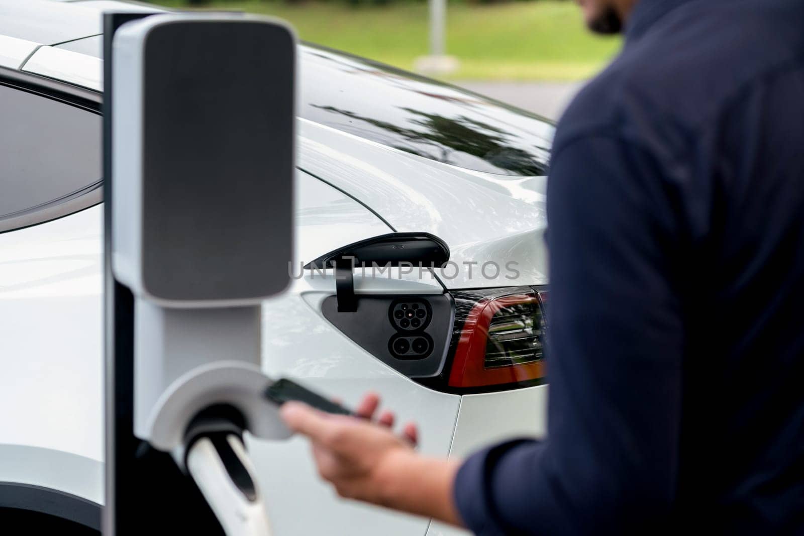 Man using smartphone online banking application to pay for electric car battery charging from EV charging station during vacation holiday road trip at national park or summer forest. Exalt