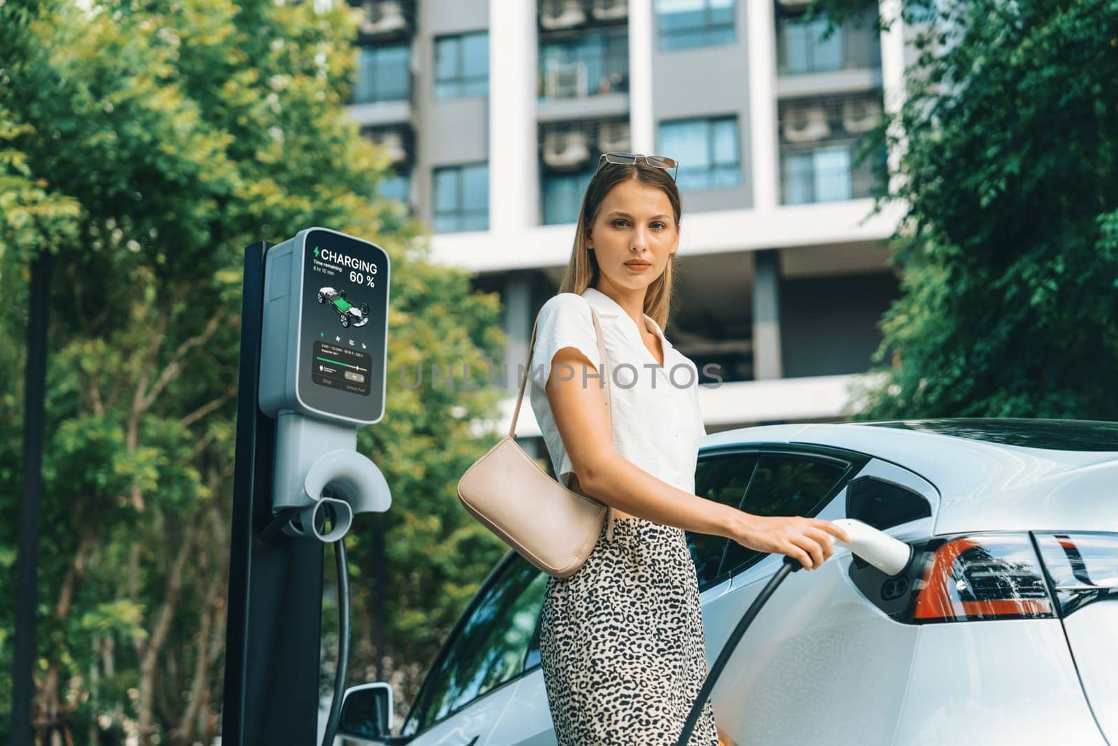 Young woman travel with EV electric car charging in green sustainable city outdoor garden in summer. Urban sustainability lifestyle by green clean rechargeable energy of electric BEV vehicle innards