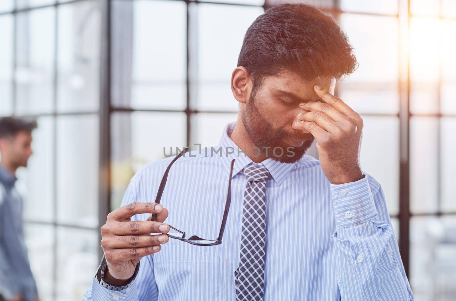 A businessman in a blue shirt and with glasses in his hand rubs his eyes from fatigue