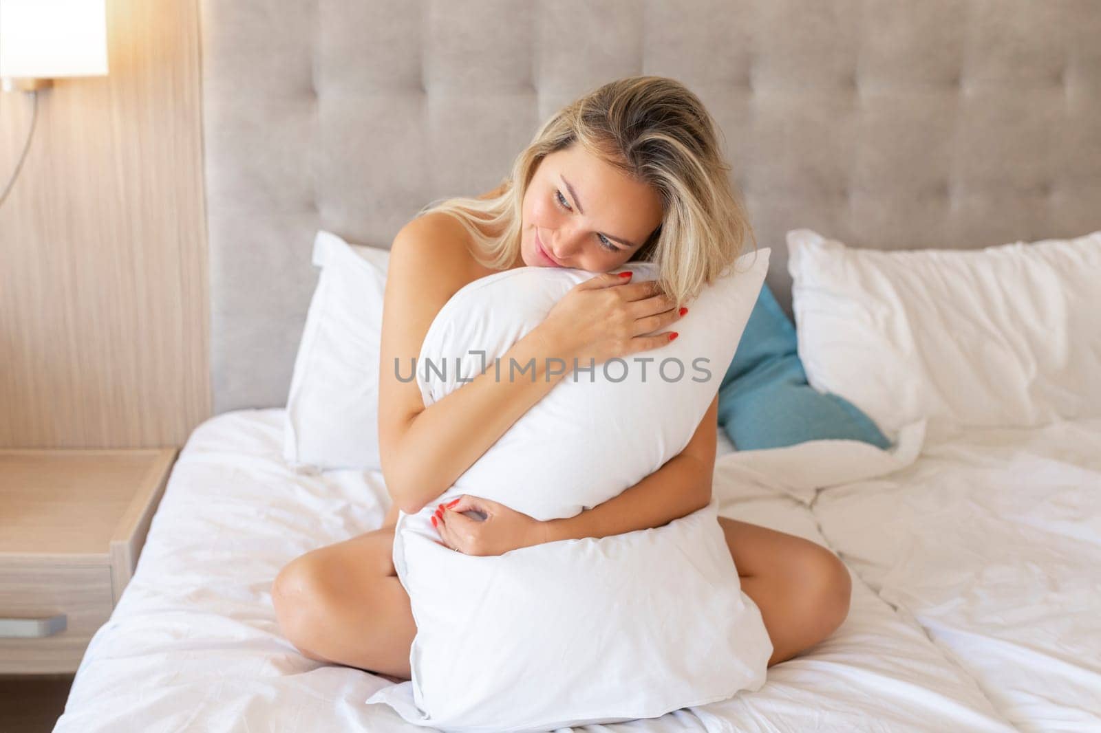 Sensual smiling young woman sitting on bed and hugging white pillow.