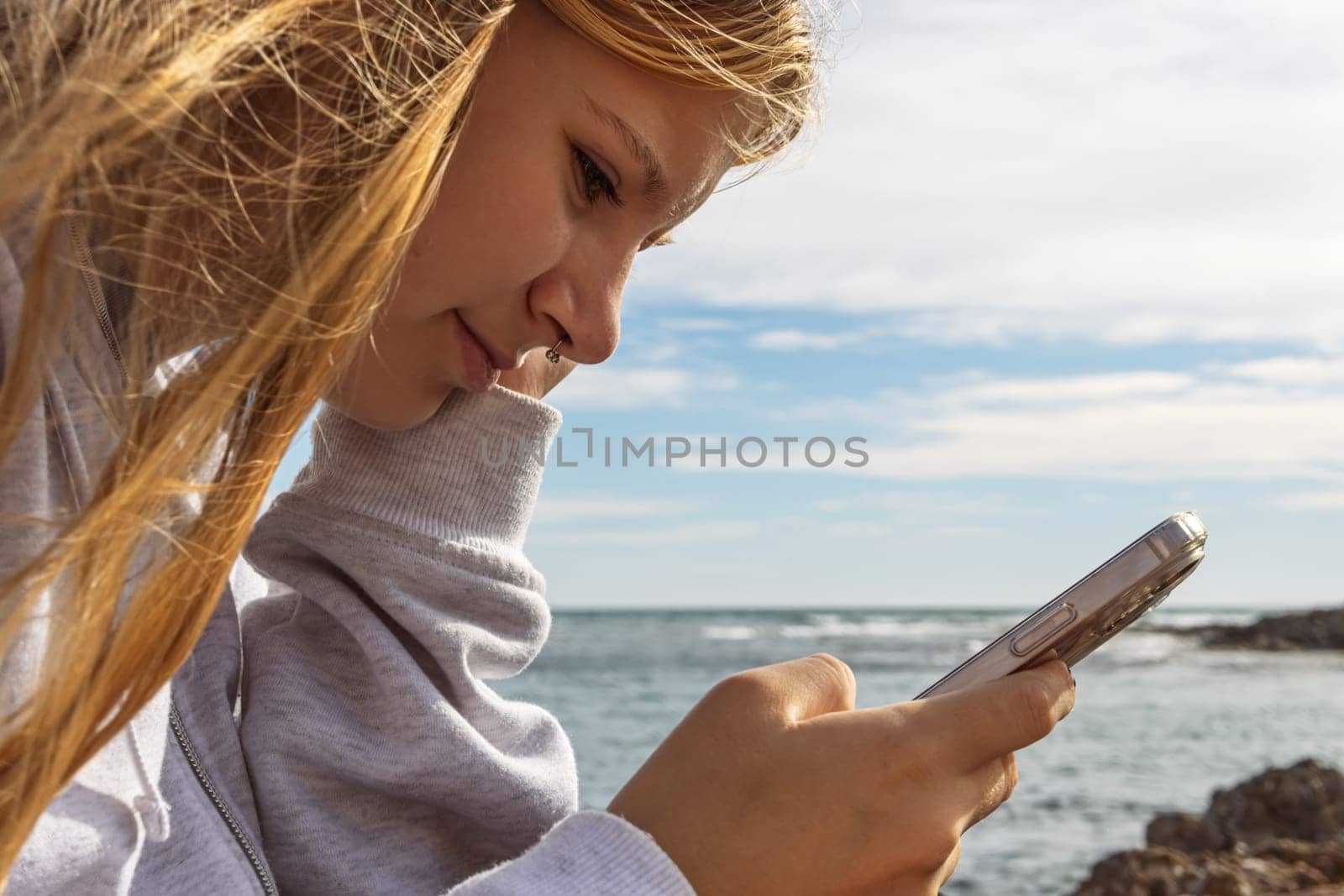 teenage girl with a piercing in the nose sitting near the sea and the girl looks at the phone by PopOff