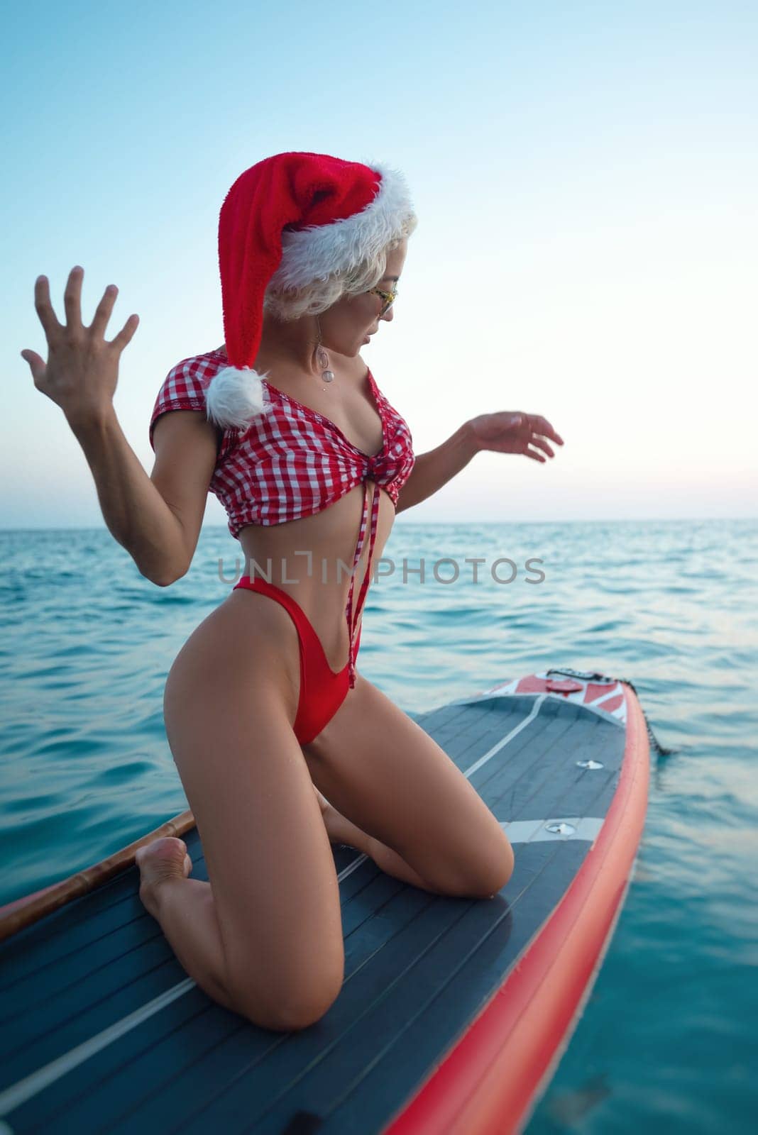 A sexy girl in a Santa Claus hat floats on the sea on a Sup board relaxes and meditates enjoys life and relaxation at sea by Rotozey