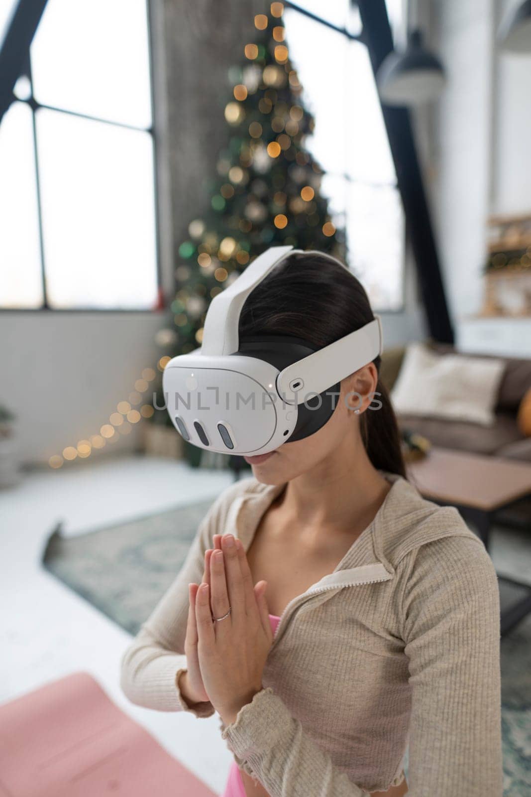 Using a virtual reality headset, a young lady in pink sportswear practices yoga near a Christmas tree. by teksomolika