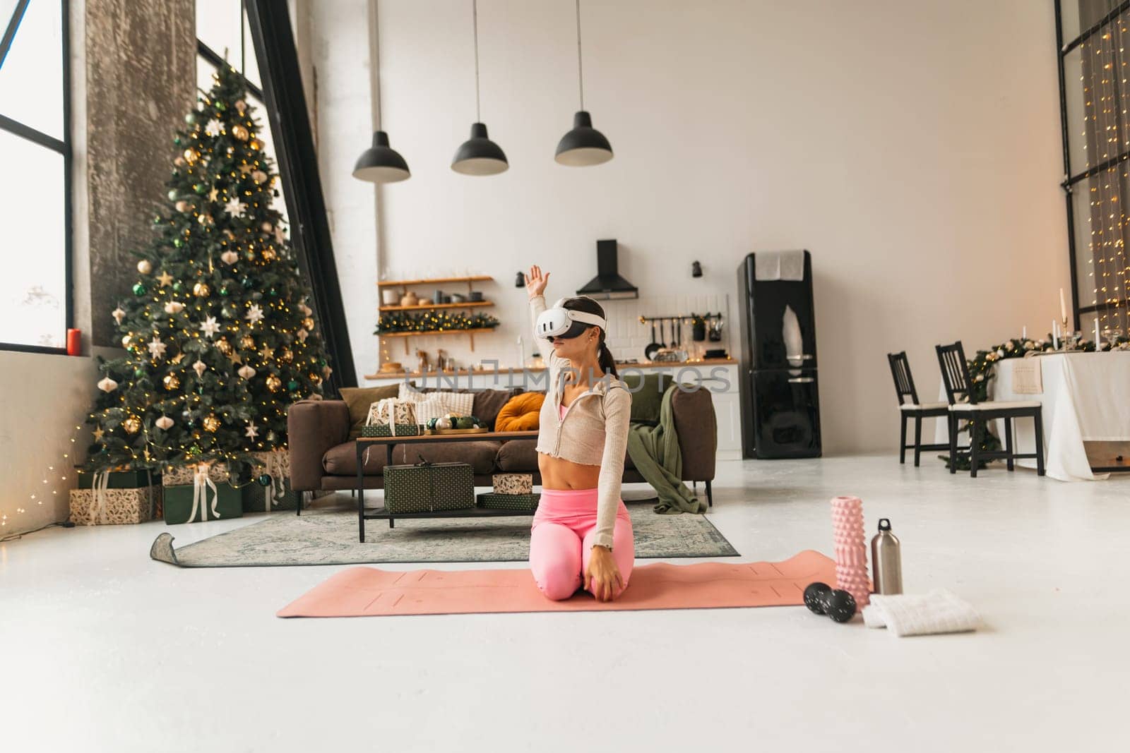 A charming young lady is doing some stretching near a Christmas tree wearing virtual reality glasses. High quality photo