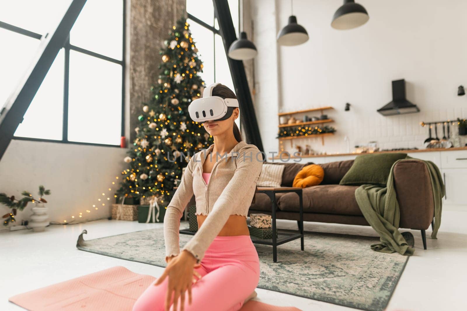 A young woman in pink sportswear practices yoga in a virtual reality headset against the backdrop of a Christmas tree. High quality photo