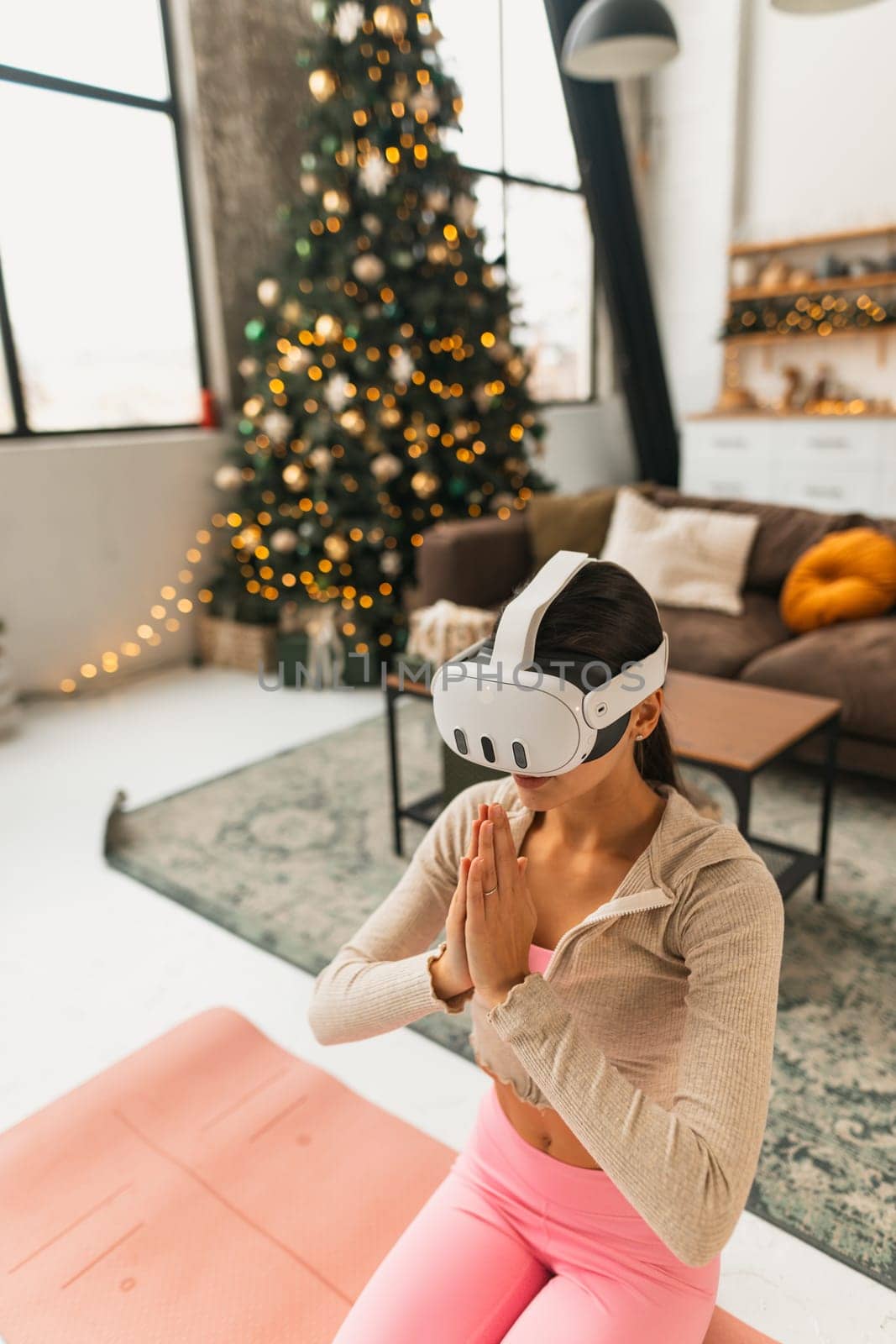 Using a virtual reality headset, a young lady in pink sportswear practices yoga near a Christmas tree. by teksomolika