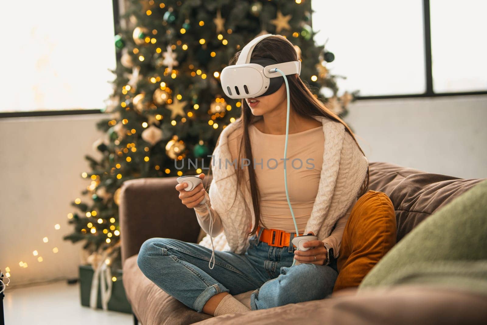 In the snug setting of a Christmas celebration at home, a fashionable young lady utilizes a virtual reality headset. by teksomolika
