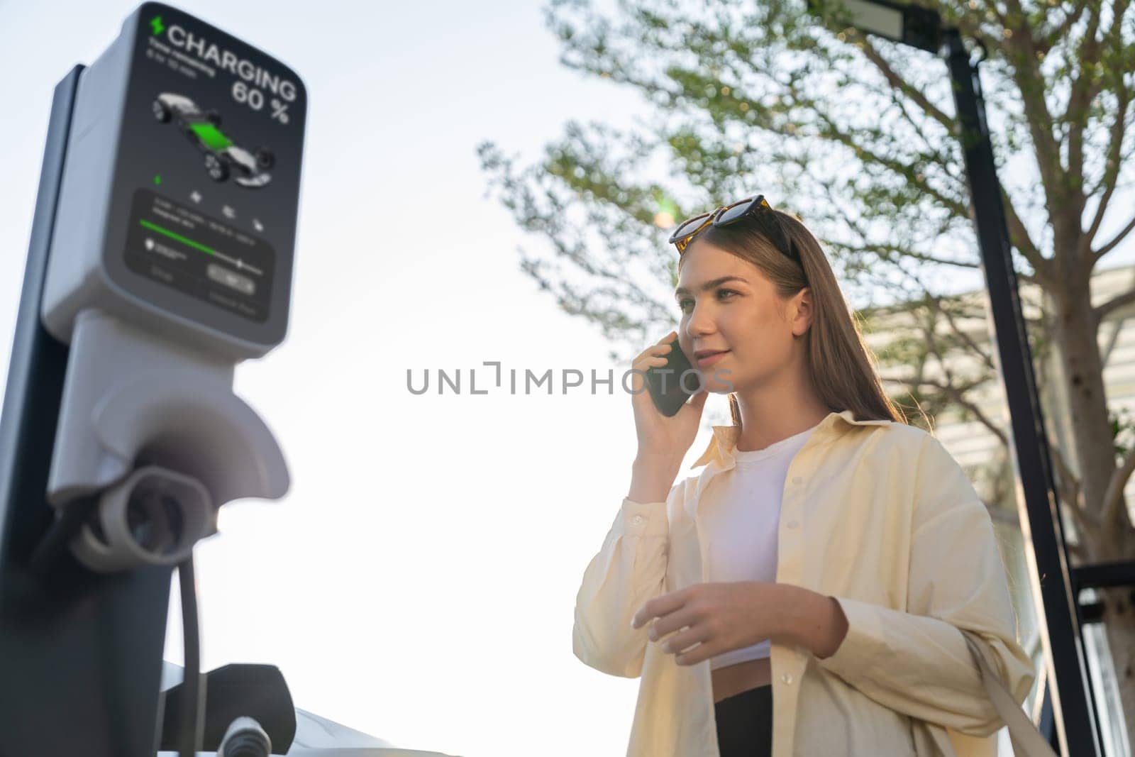 Young woman holding shopping bag talking on the phone while recharging EV car battery from charging station at city mall parking lot. Modern woman go shopping by eco car. Expedient