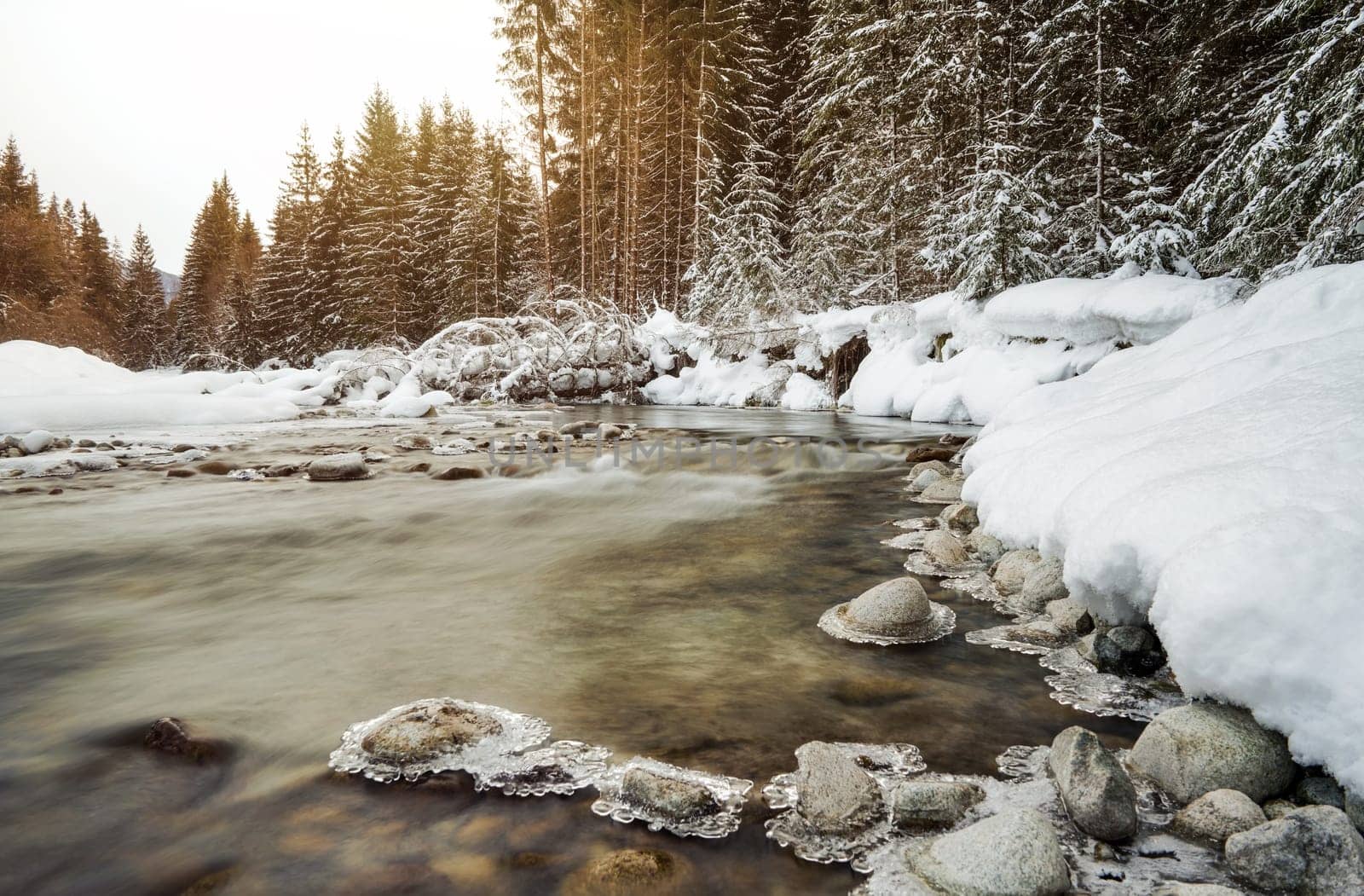 Morning sun shines on forest river creek in winter, rocks on shore covered with snow, long exposure makes water silky smooth