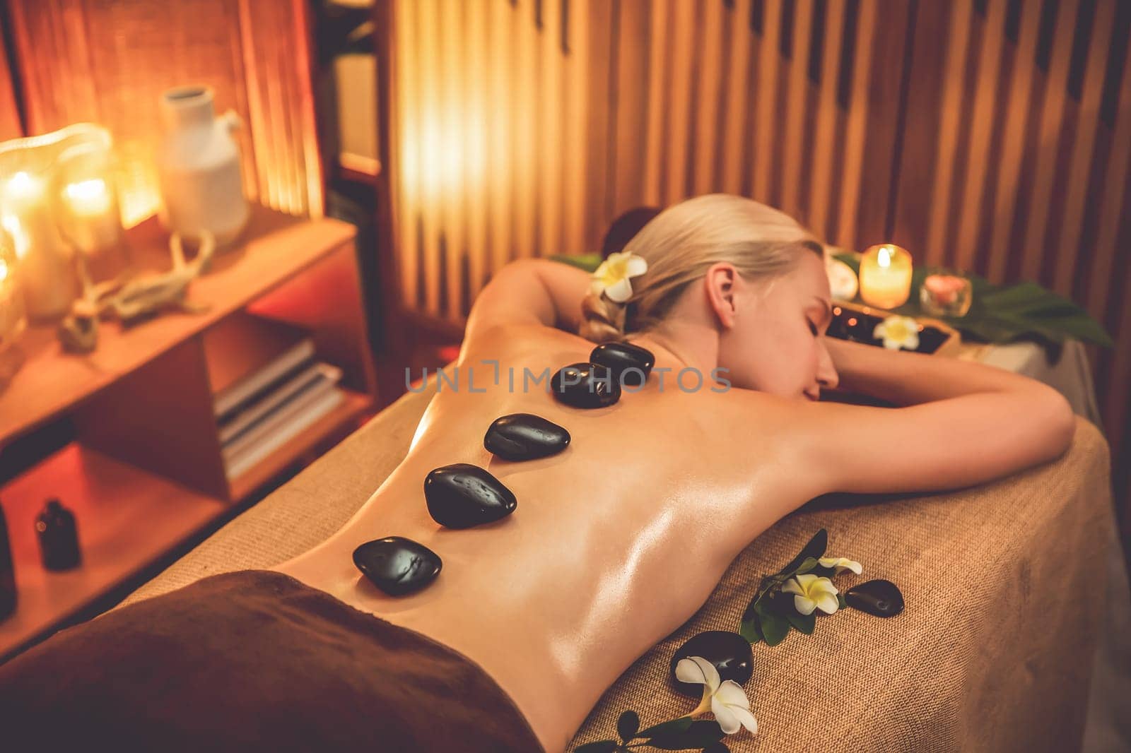 Hot stone massage at spa salon in luxury resort with warm candle light, blissful woman customer enjoying spa basalt stone massage glide over body with soothing warmth. Quiescent