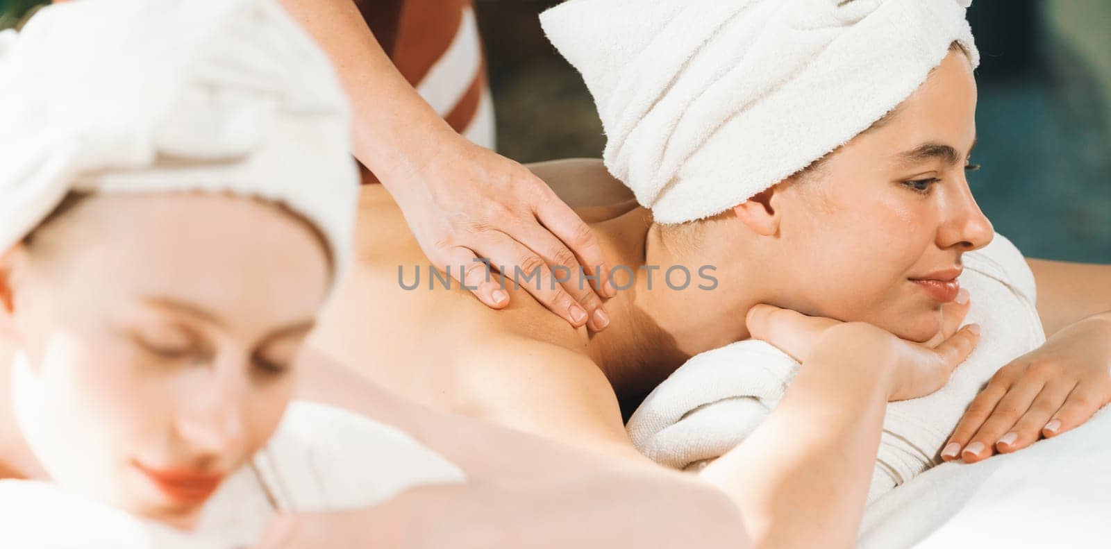 A couple of beautiful young woman with white towel lie on comfortable spa bed during receive relaxing body massage by professional masseur surrounded at spa salon. Gray background. Tranquility.