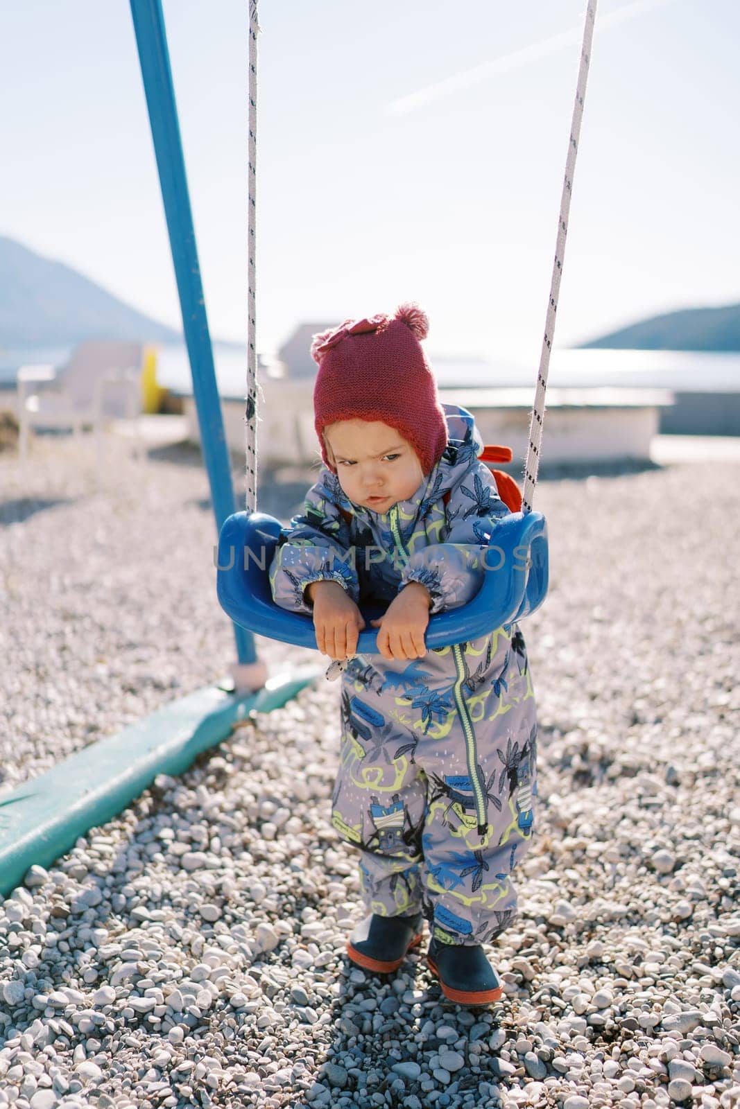 Little girl stands leaning on the seat of a rope swing in the playground. High quality photo