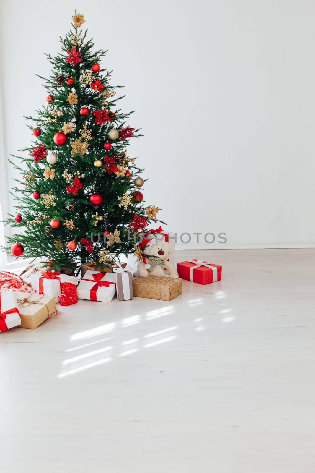 Big beautiful christmas tree decorated with beautiful shiny baubles and many different presents on wooden floor. White wall background with a lot of copy space for text. Close up.