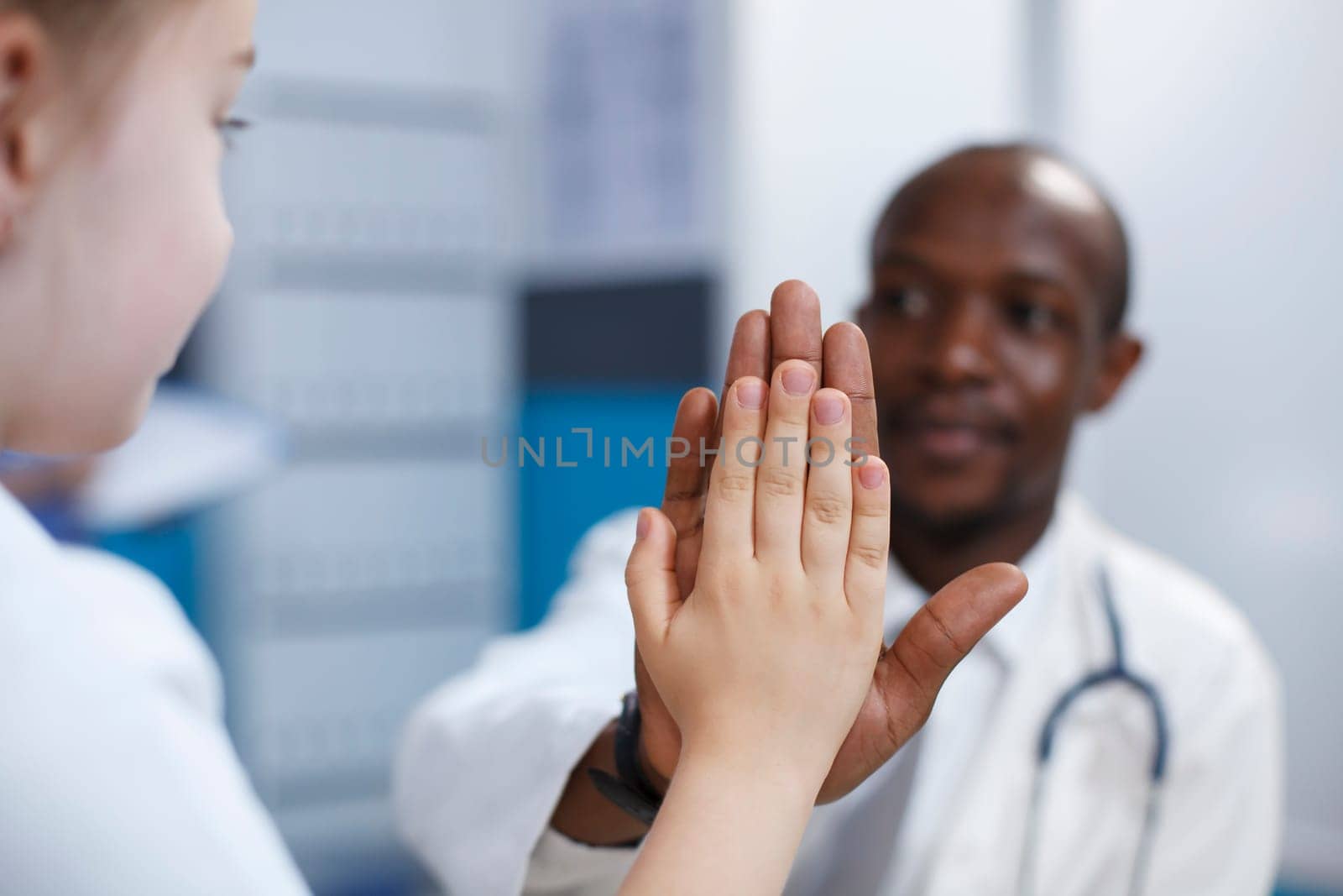 Selective focus of a caucasian girl high fiving with an african american male pediatrician in a hospital office. Close-up shot of friendly doctor having medical consultation with a female youngster.