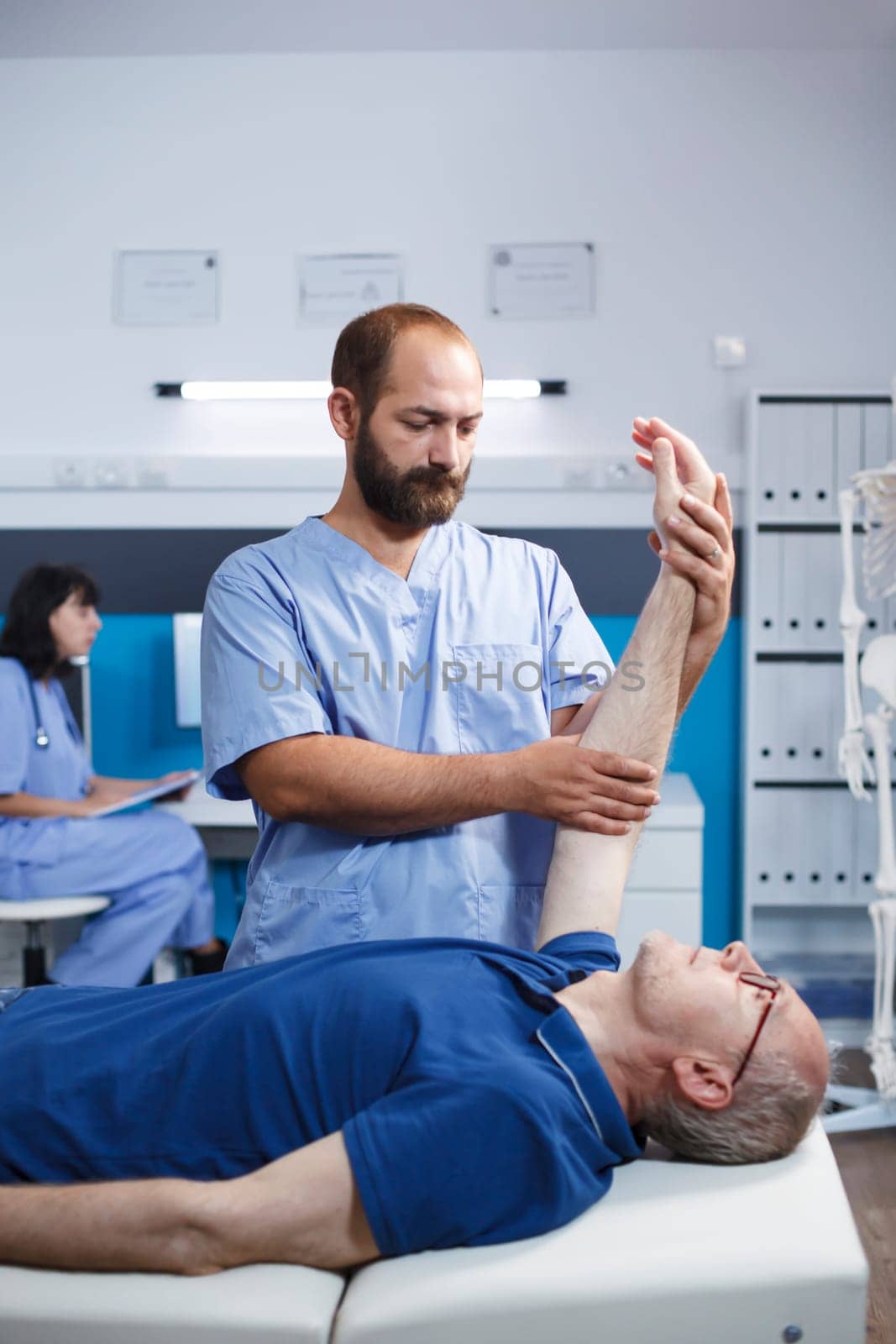 Male elderly patient receiving osteopathic care from a medical practitioner for arm pain. Caucasian retired old man aided by nurse in blue scrubs through physiotherapy exercise.