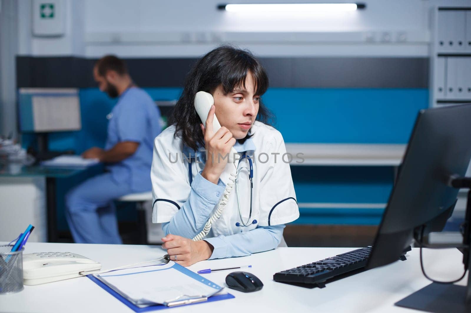 Caucasian doctor in modern office sitting at desk, speaking on landline. With a desktop PC on the table, a female physician is making a phone call. Technology and communication in healthcare.