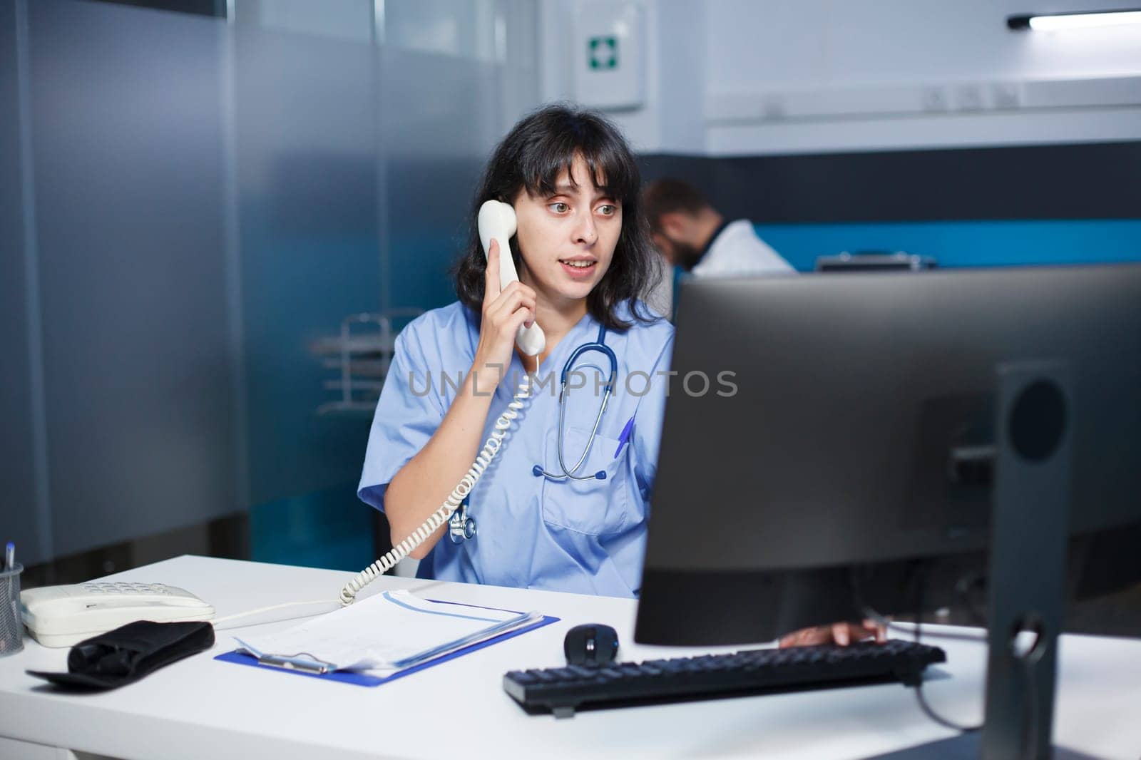 In the hospital office, a medical practitioner answers phones and schedules appointments. In a modern clinic, a health care provider sits at a desk, using a landline and staring at a monitor.