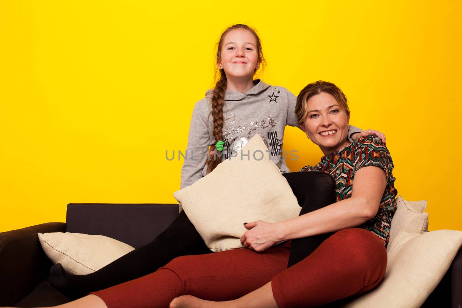 cheerful girl with mom at home on black sofa at yellow wall