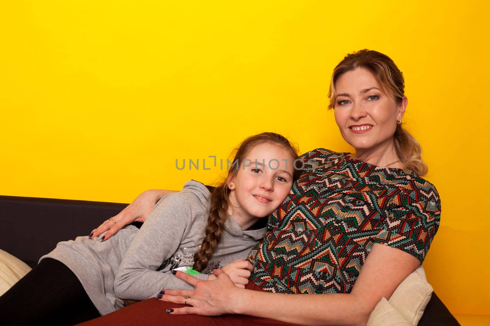 cheerful girl with mom at home on black sofa at yellow wall