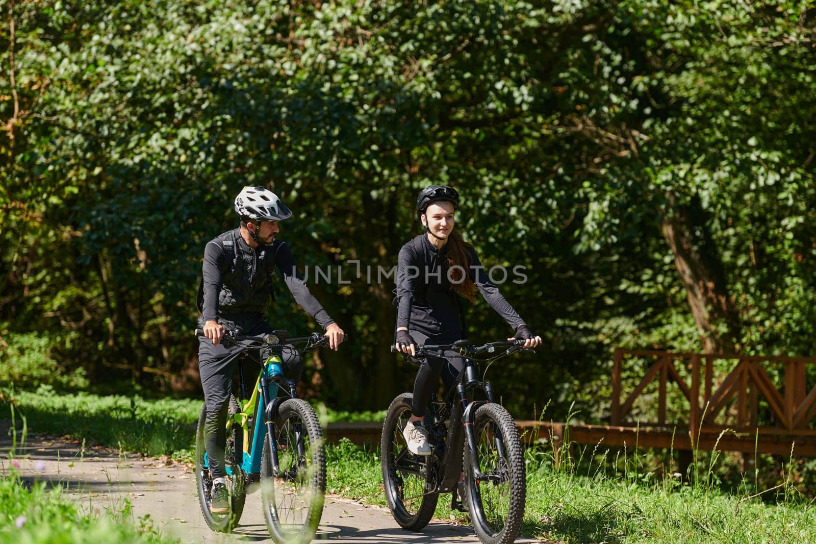 A blissful couple, adorned in professional cycling gear, enjoys a romantic bicycle ride through a park, surrounded by modern natural attractions, radiating love and happiness by dotshock