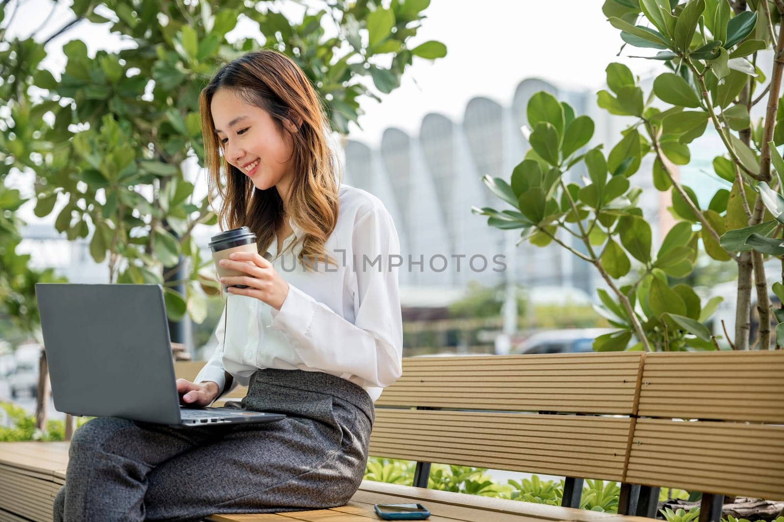 Happy Asian professional smiling businesswoman sitting alone typing computer outside street city near office, Successful business young woman working laptop outdoor corporate building exterior