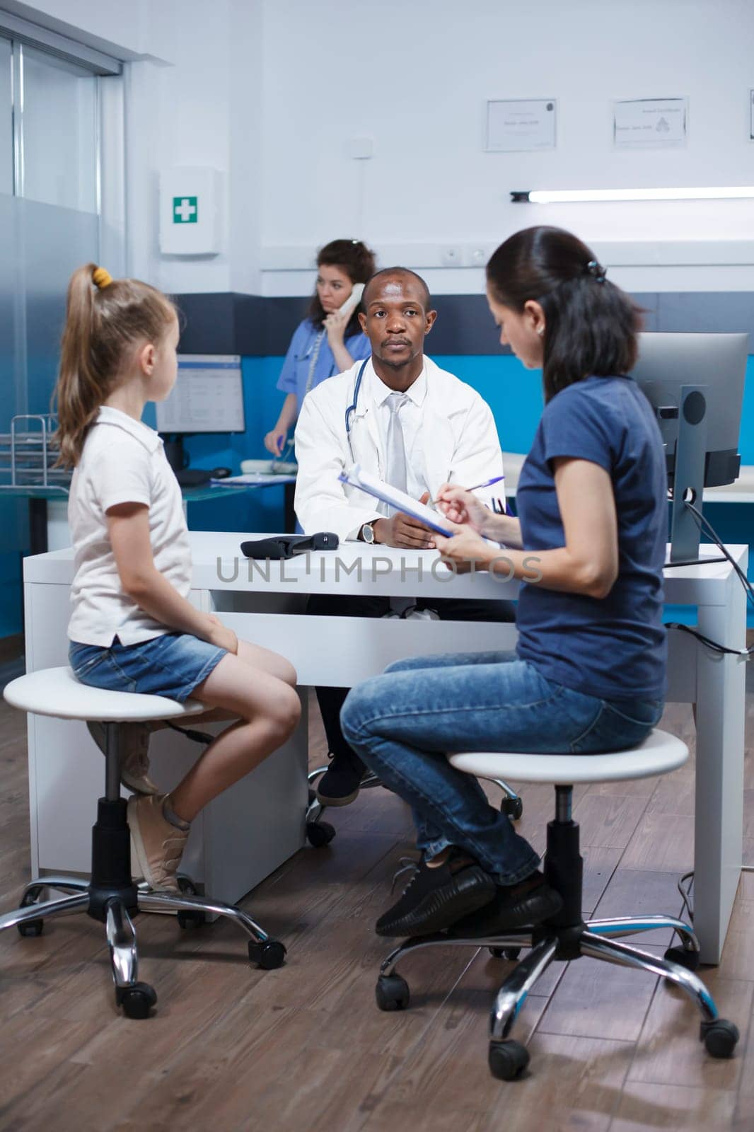 Caucasian woman in the hospital office with her daughter filling out the papers for a medical examination. African American doctor waits as the lady completes paperwork at the clinic.