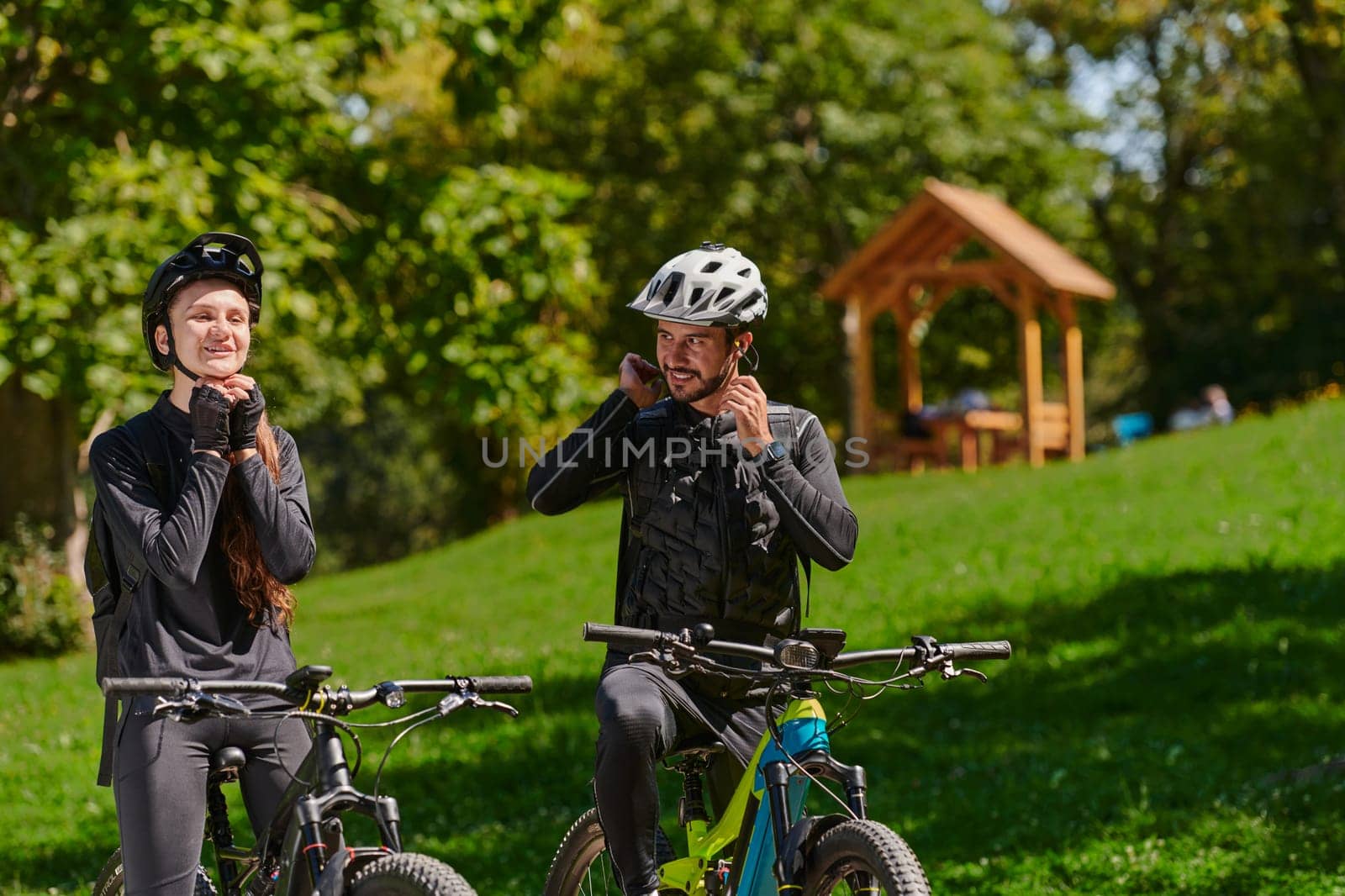 A sweet couple, equipped with bicycles and engrossed in coordinating their journey, checks their GPS mobile and watches while planning scenic routes in the park, seamlessly blending technology and outdoor adventure for a delightful cycling experience by dotshock