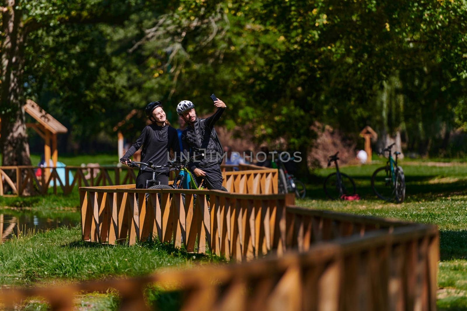 A blissful couple, adorned in professional cycling gear, enjoys a romantic bicycle ride through a park, surrounded by modern natural attractions, radiating love and happiness.