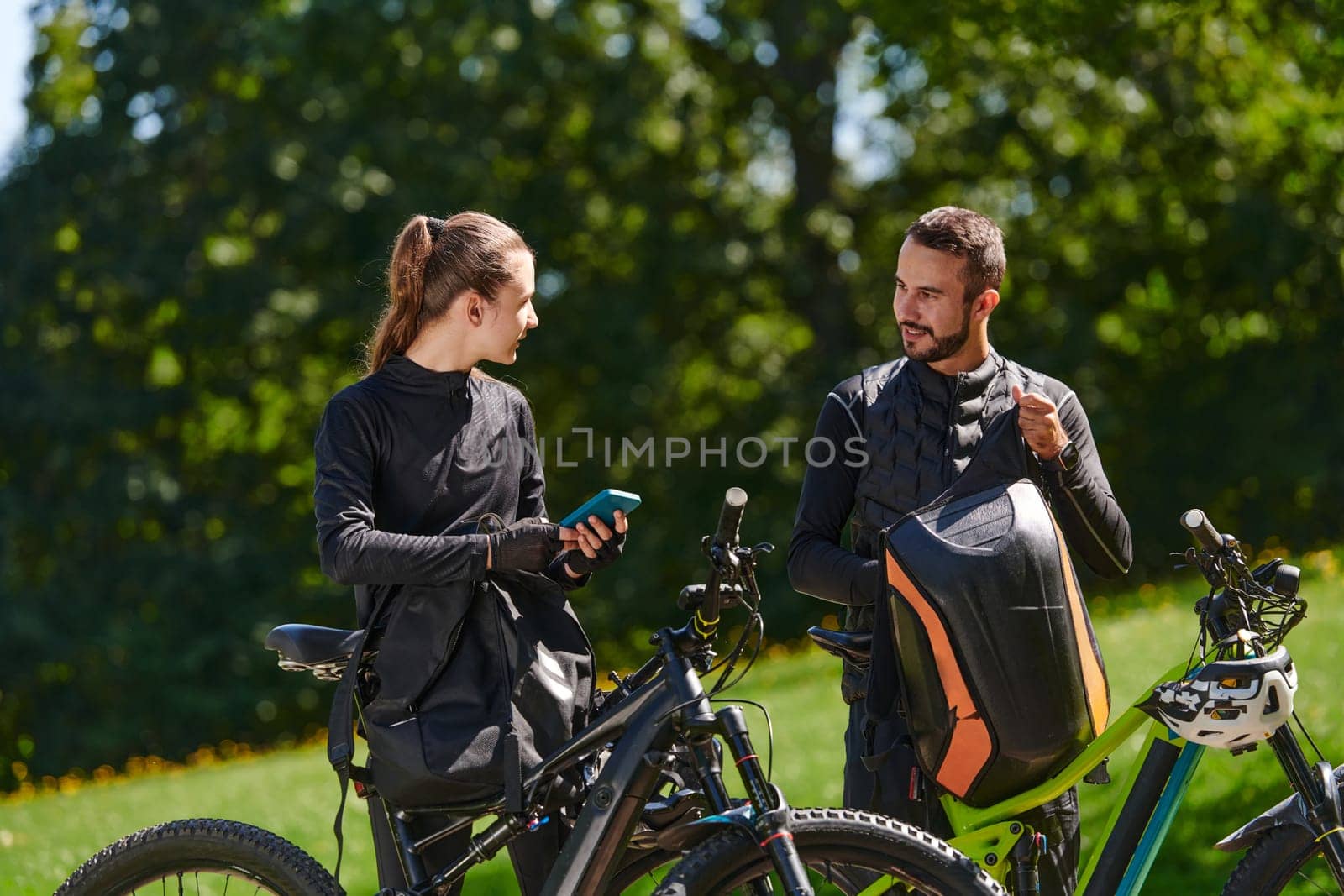 A sweet couple, equipped with bicycles and engrossed in coordinating their journey, checks their GPS mobile and watches while planning scenic routes in the park, seamlessly blending technology and outdoor adventure for a delightful cycling experience.