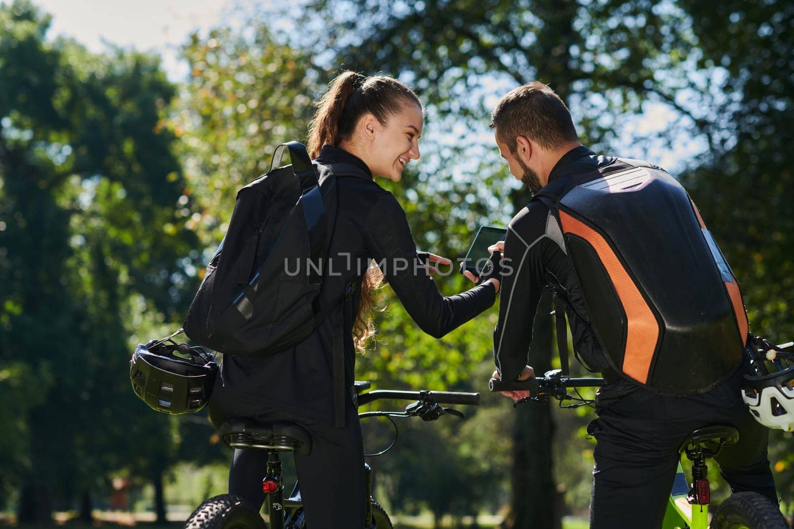 A sweet couple, equipped with bicycles and engrossed in coordinating their journey, checks their GPS mobile and watches while planning scenic routes in the park, seamlessly blending technology and outdoor adventure for a delightful cycling experience by dotshock