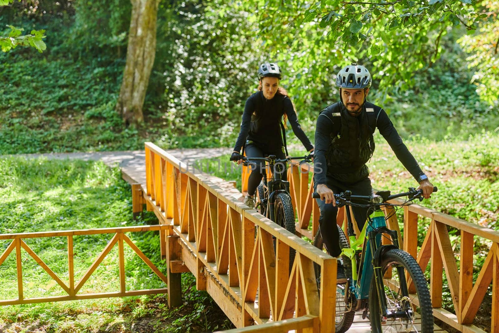 A blissful couple, adorned in professional cycling gear, enjoys a romantic bicycle ride through a park, surrounded by modern natural attractions, radiating love and happiness by dotshock