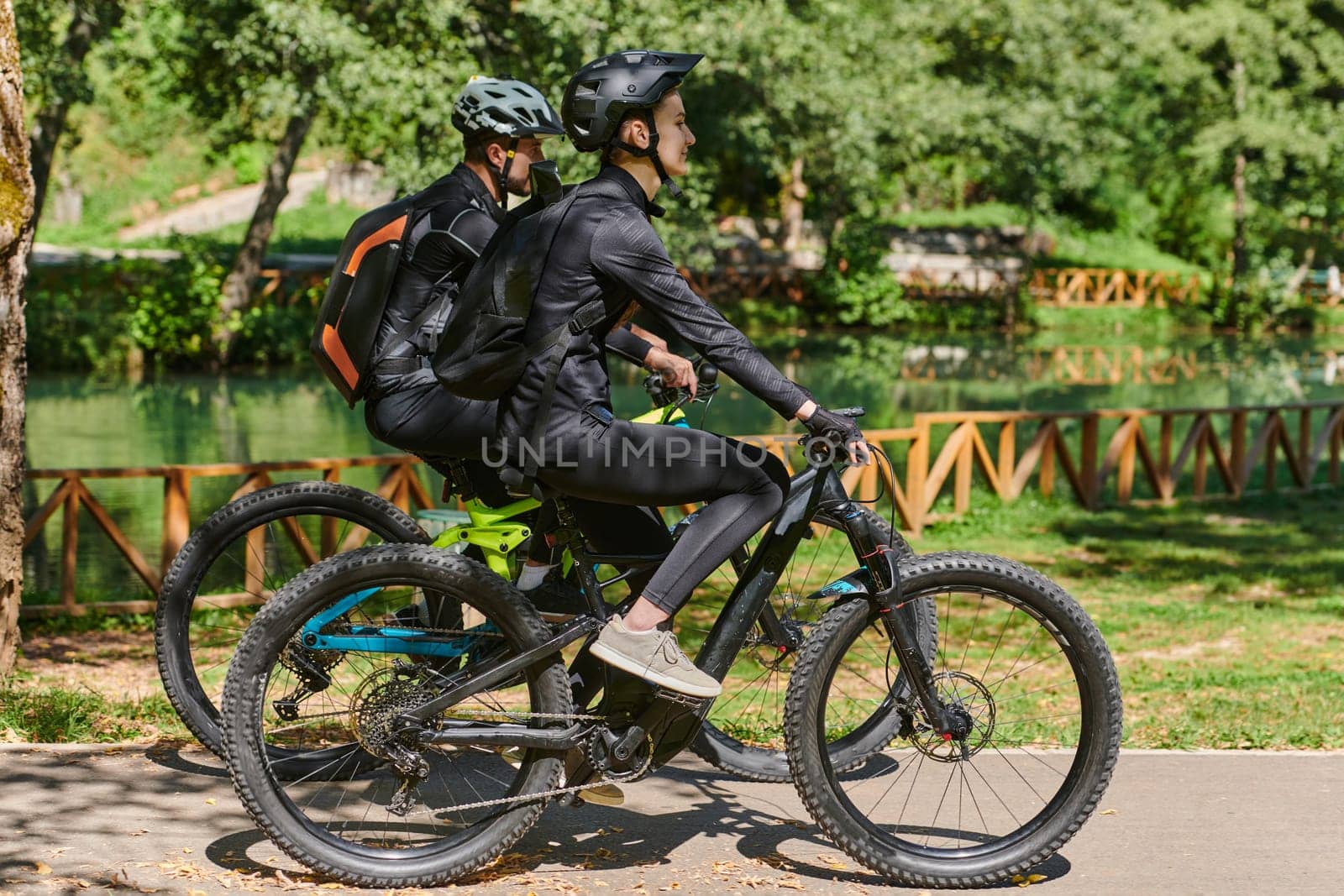 A blissful couple, adorned in professional cycling gear, enjoys a romantic bicycle ride through a park, surrounded by modern natural attractions, radiating love and happiness by dotshock