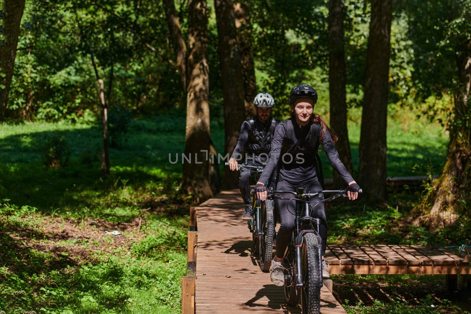 A blissful couple, adorned in professional cycling gear, enjoys a romantic bicycle ride through a park, surrounded by modern natural attractions, radiating love and happiness.