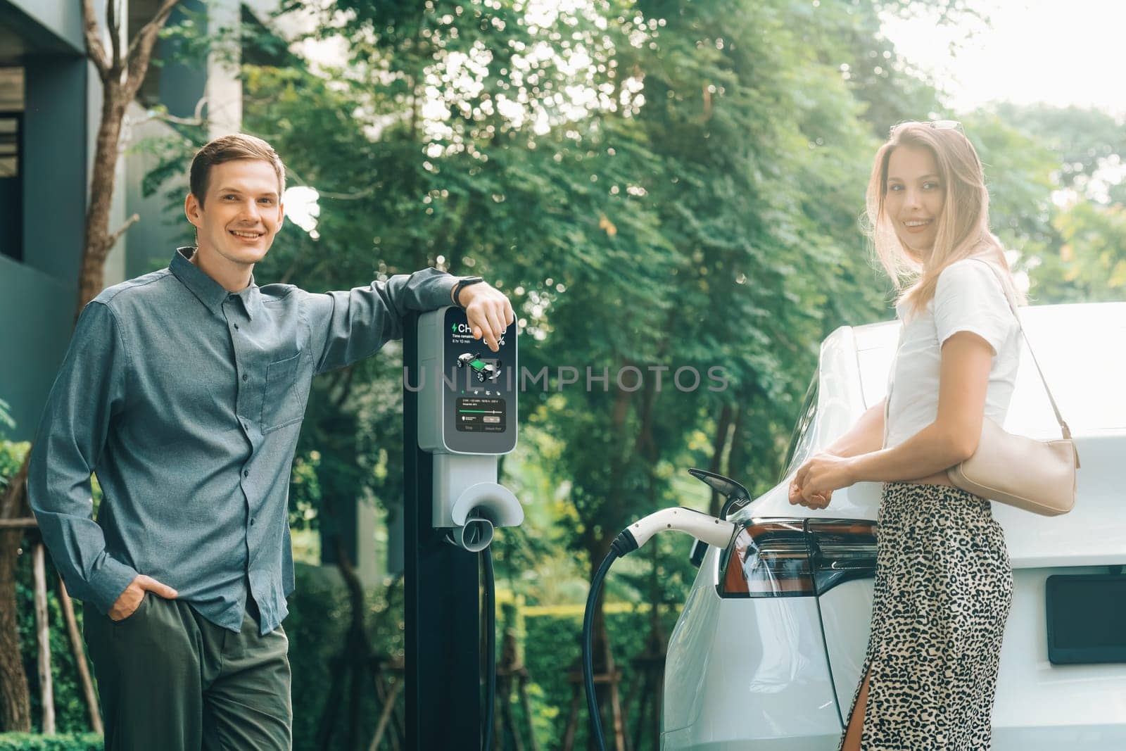 Young couple travel with EV electric car charging in green sustainable city outdoor garden in summer shows urban sustainability lifestyle by green clean rechargeable energy of electric vehicle innards