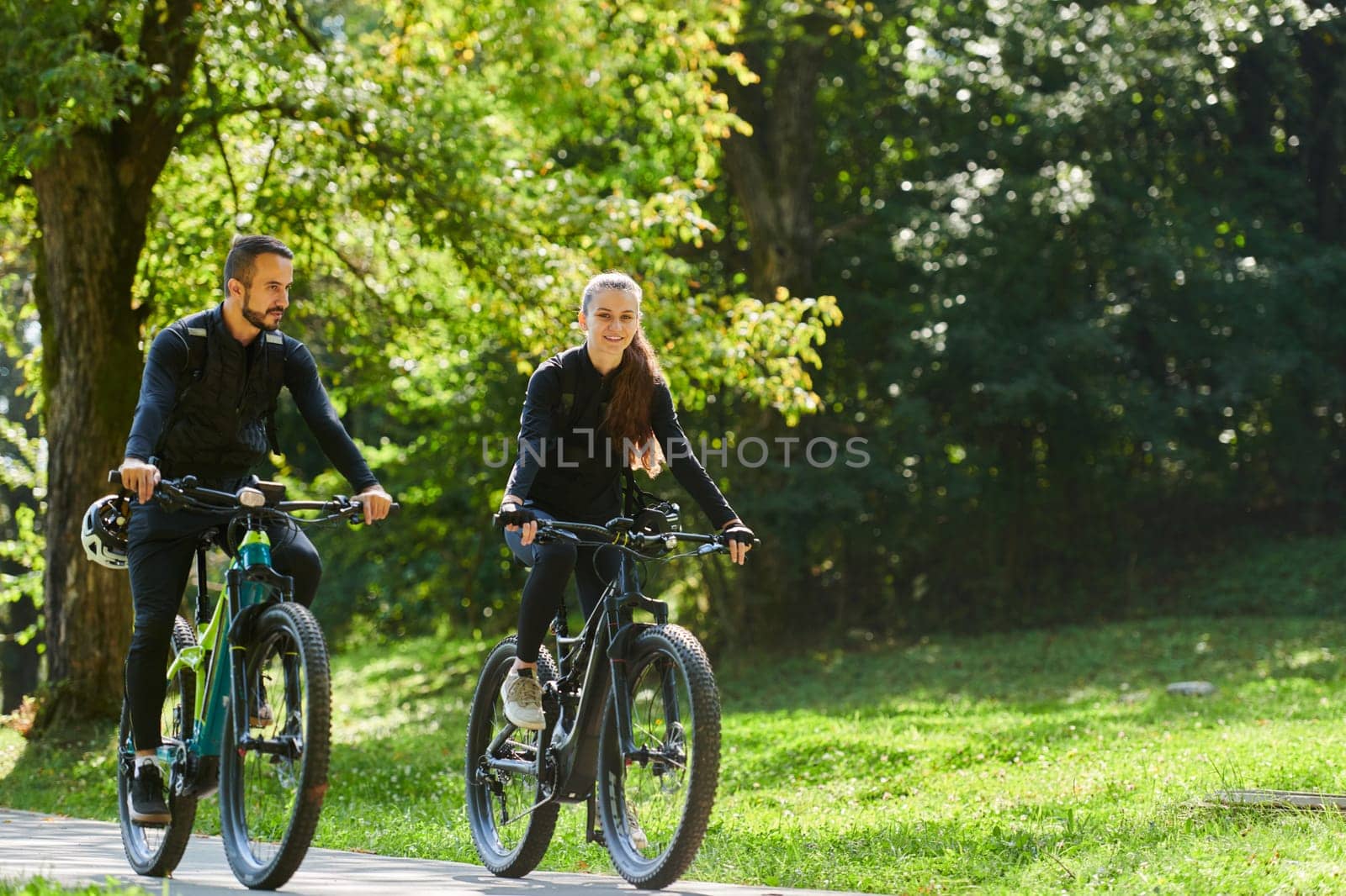 A blissful couple, adorned in professional cycling gear, enjoys a romantic bicycle ride through a park, surrounded by modern natural attractions, radiating love and happiness.