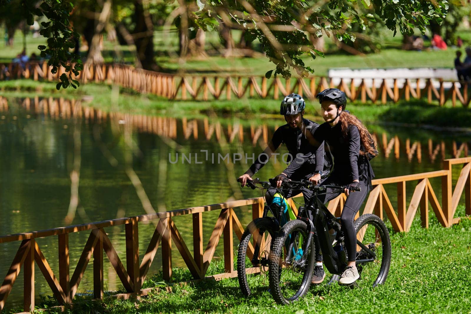 A blissful couple, adorned in professional cycling gear, enjoys a romantic bicycle ride through a park, surrounded by modern natural attractions, radiating love and happiness by dotshock