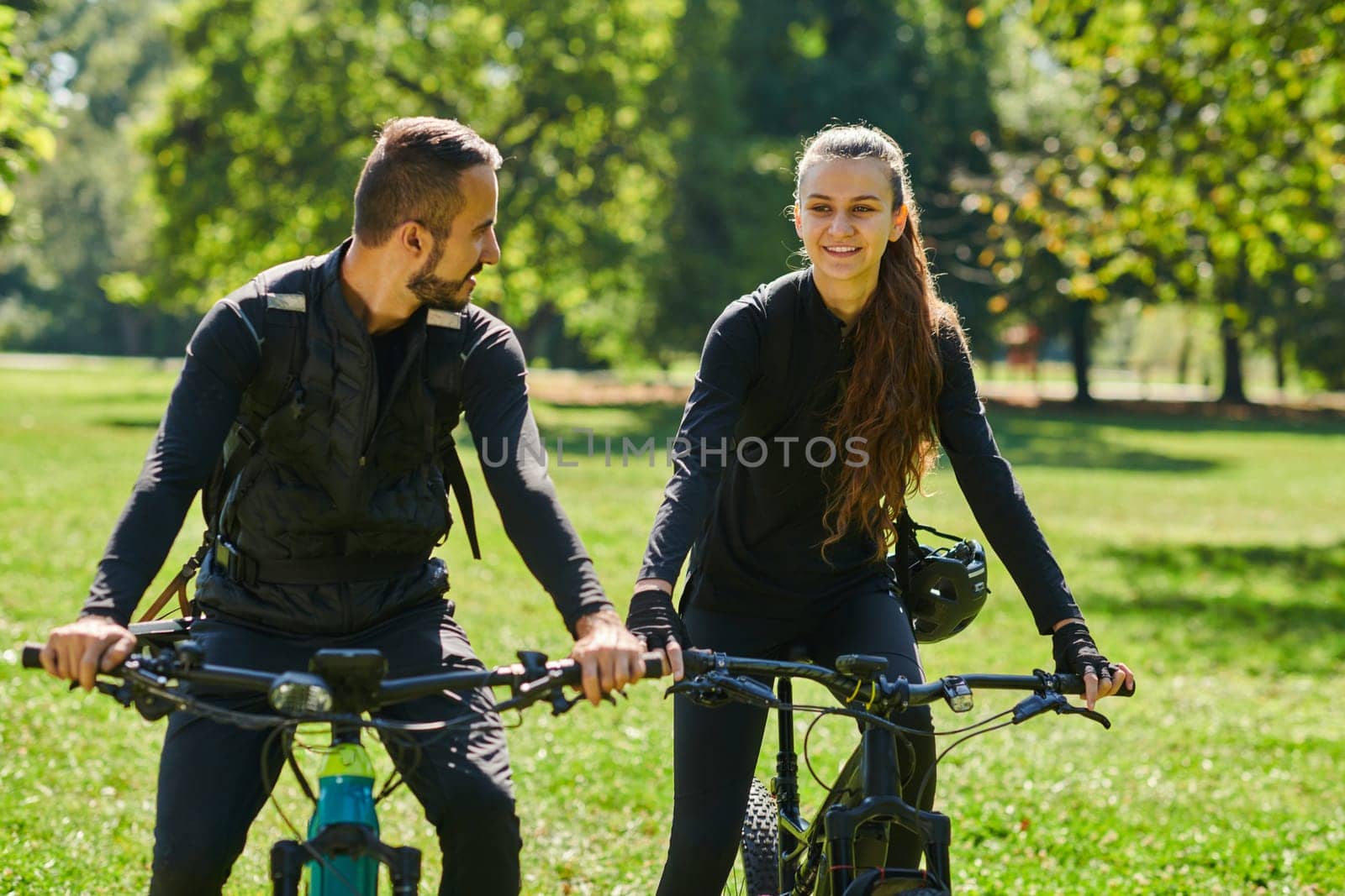 A blissful couple, adorned in professional cycling gear, enjoys a romantic bicycle ride through a park, surrounded by modern natural attractions, radiating love and happiness.