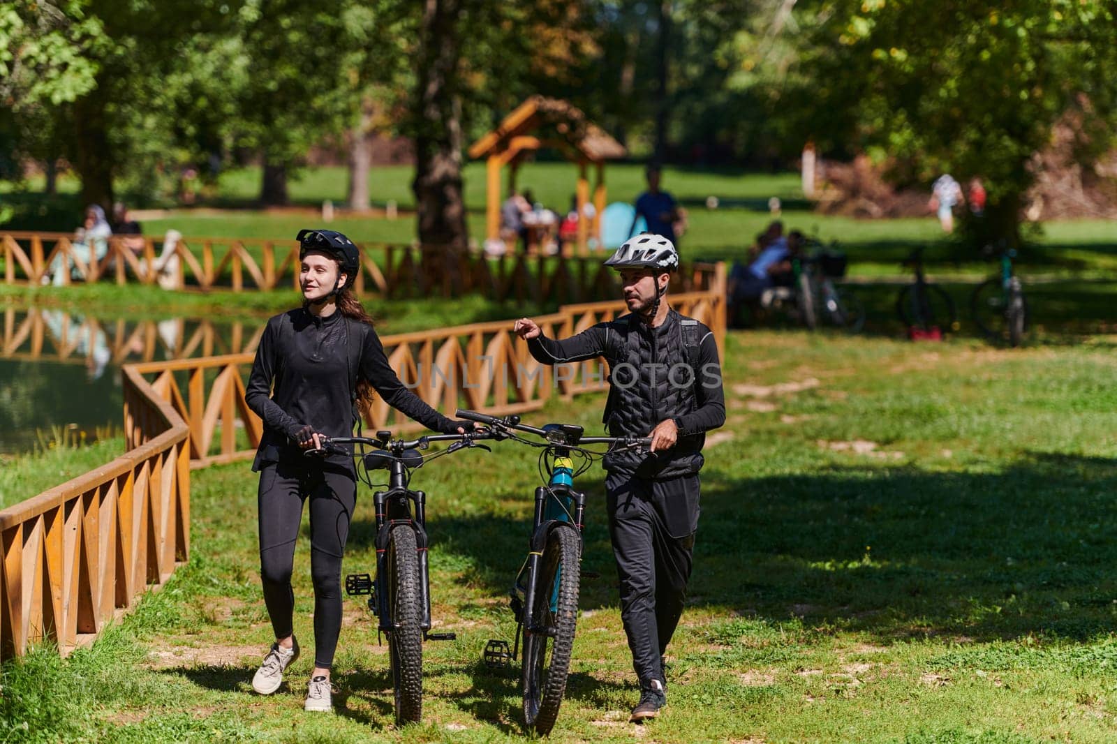 A blissful couple, adorned in professional cycling gear, enjoys a romantic bicycle ride through a park, surrounded by modern natural attractions, radiating love and happiness by dotshock