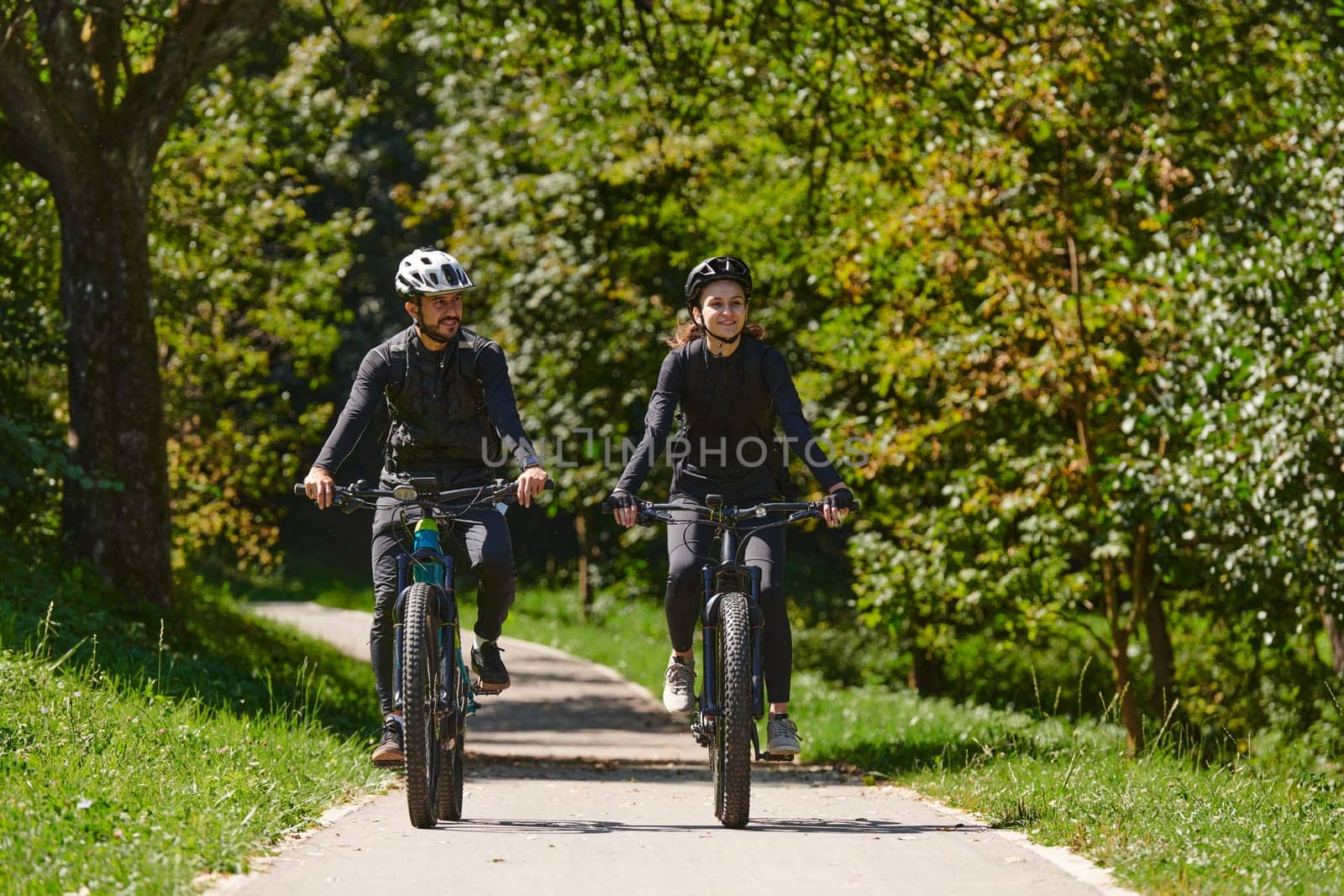 A blissful couple, adorned in professional cycling gear, enjoys a romantic bicycle ride through a park, surrounded by modern natural attractions, radiating love and happiness by dotshock