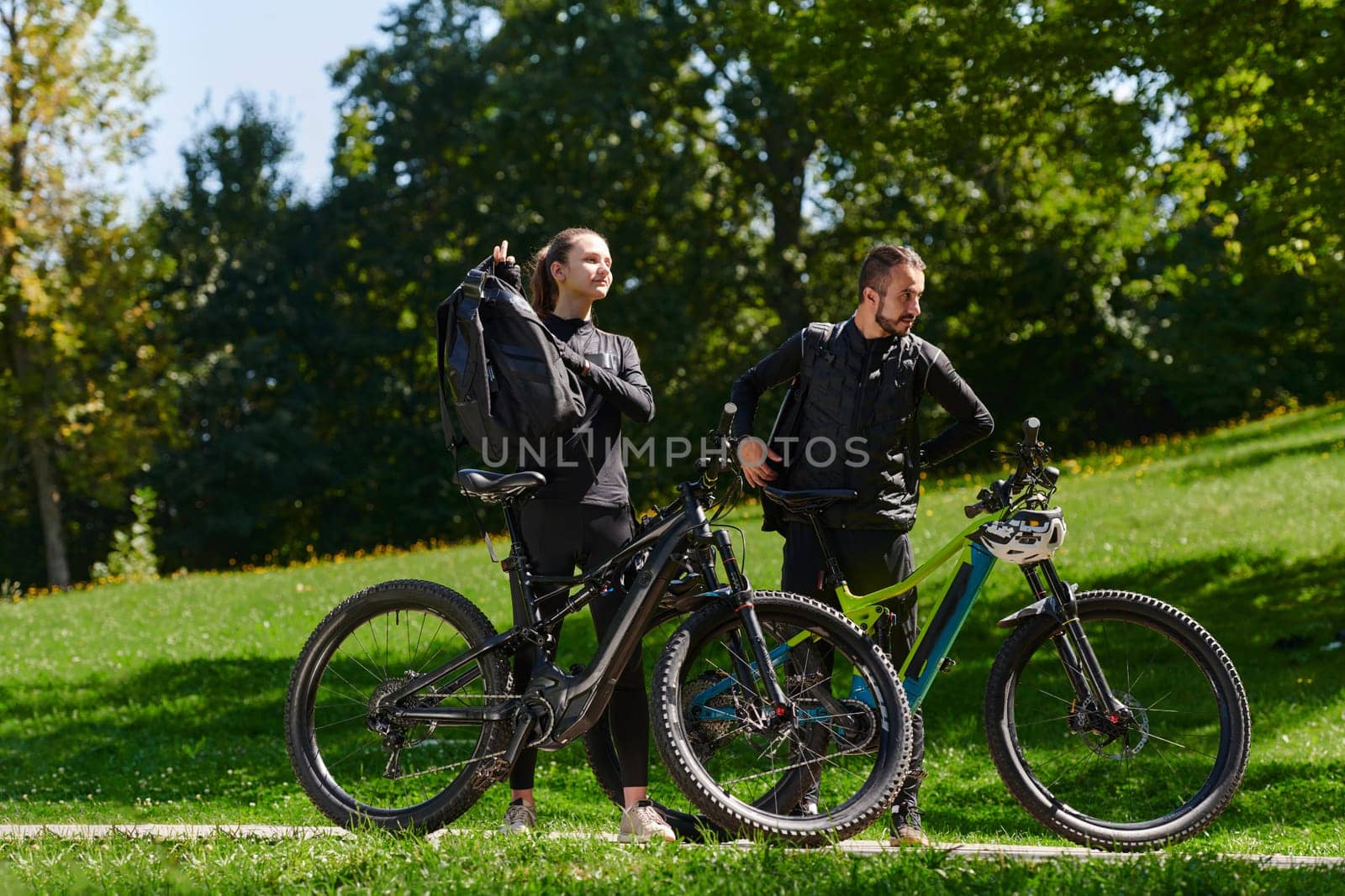 A sweet couple, equipped with bicycles and engrossed in coordinating their journey, checks their GPS mobile and watches while planning scenic routes in the park, seamlessly blending technology and outdoor adventure for a delightful cycling experience.