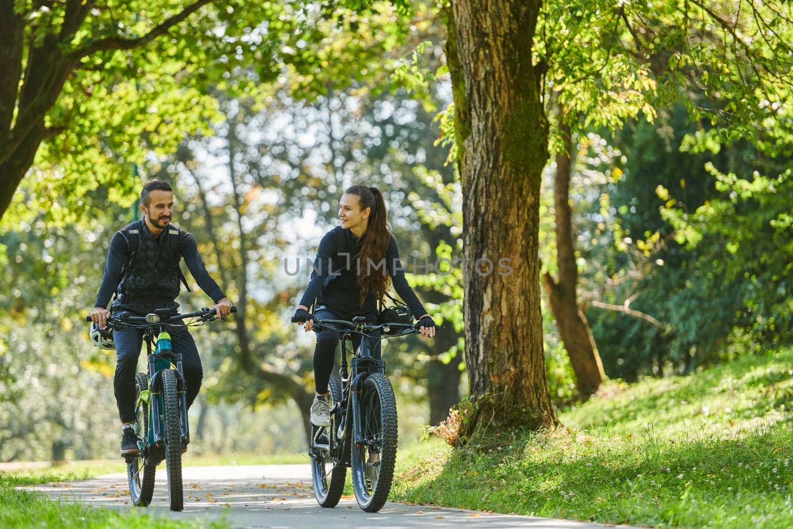 A blissful couple, adorned in professional cycling gear, enjoys a romantic bicycle ride through a park, surrounded by modern natural attractions, radiating love and happiness.