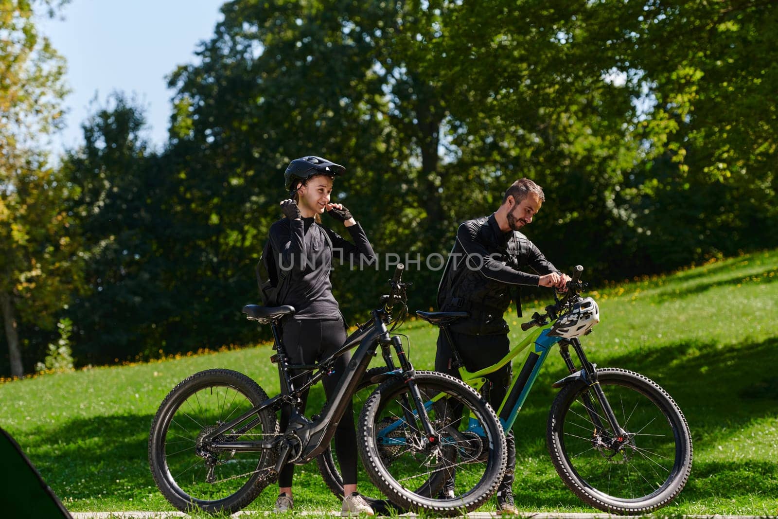 A sweet couple, equipped with bicycles and engrossed in coordinating their journey, checks their GPS mobile and watches while planning scenic routes in the park, seamlessly blending technology and outdoor adventure for a delightful cycling experience by dotshock