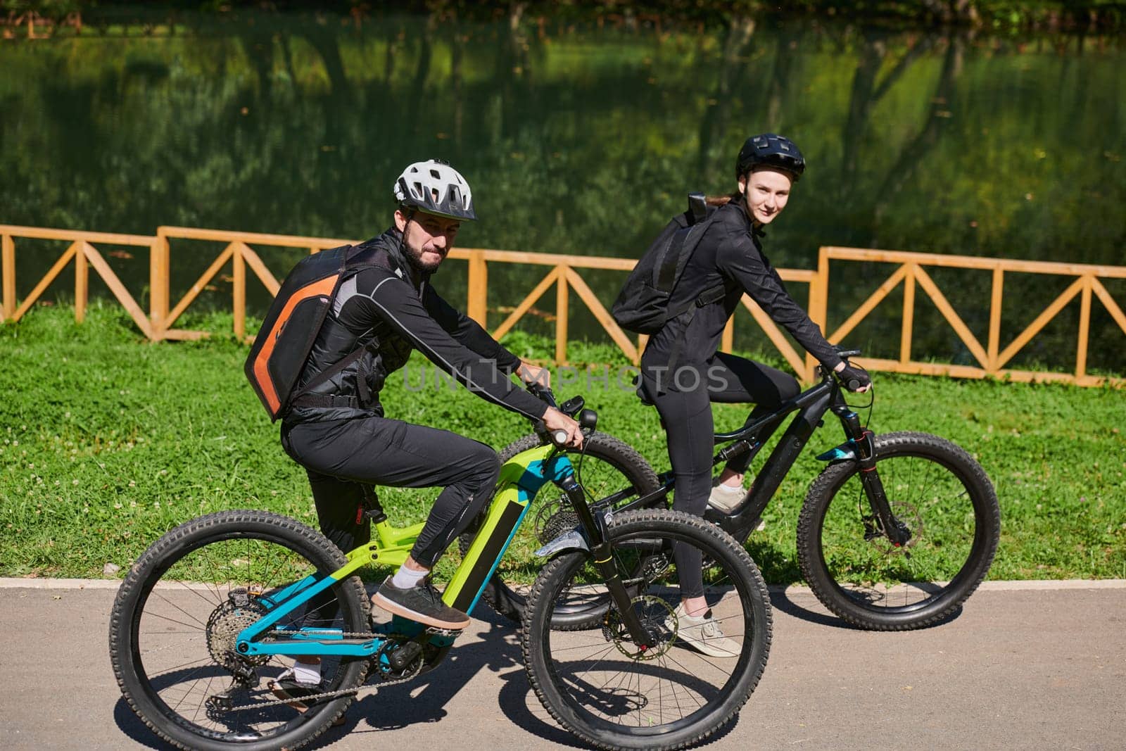 A blissful couple, adorned in professional cycling gear, enjoys a romantic bicycle ride through a park, surrounded by modern natural attractions, radiating love and happiness.